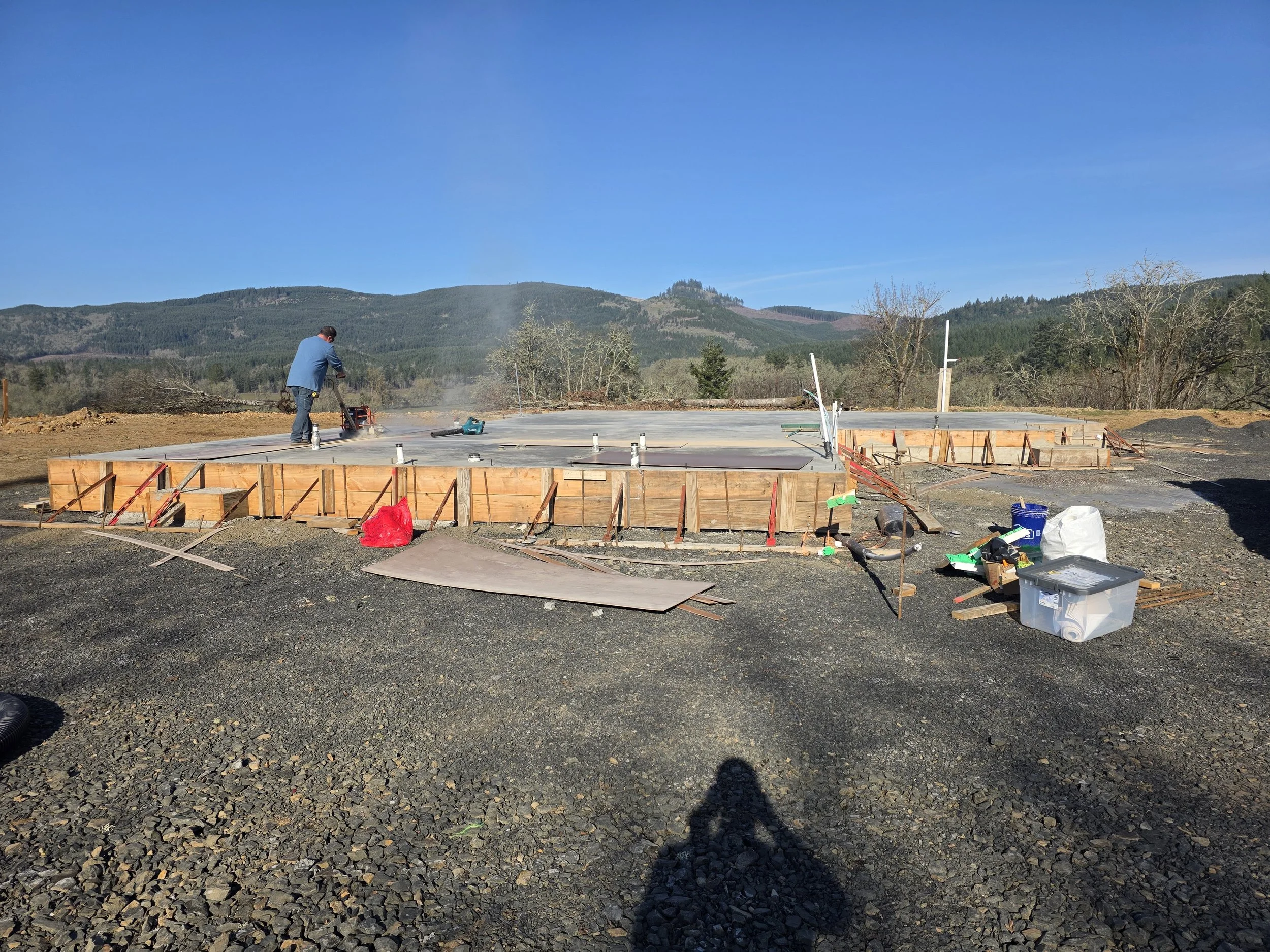 Construction site with a worker operating equipment on a concrete slab, set against a backdrop of mountains and blue sky. New house Construction Turner Oregon.