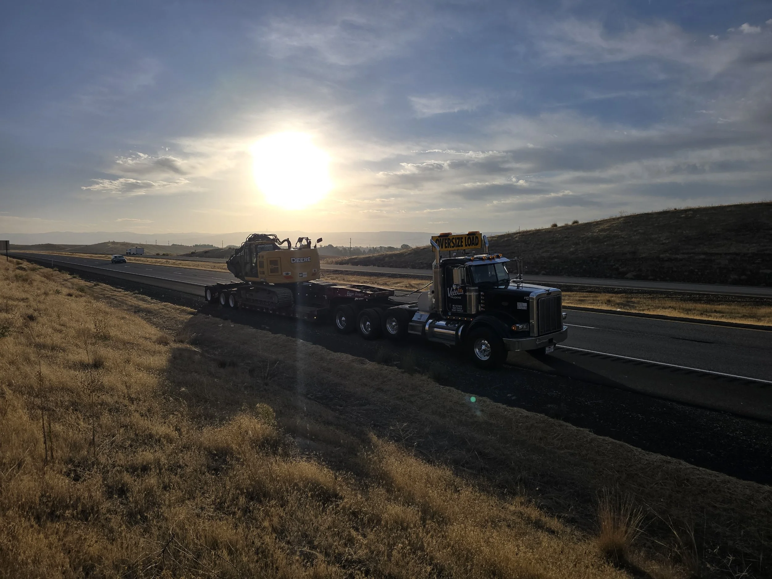 A truck with an oversized load sign is transporting construction equipment, including a bulldozer, along a highway during sunset.