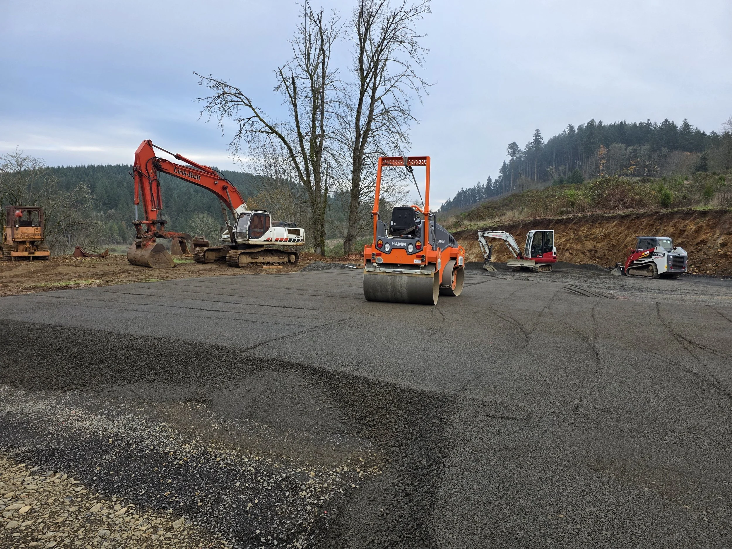 Construction site with compacting equipment and excavators on a dirt area surrounded by trees and hills.