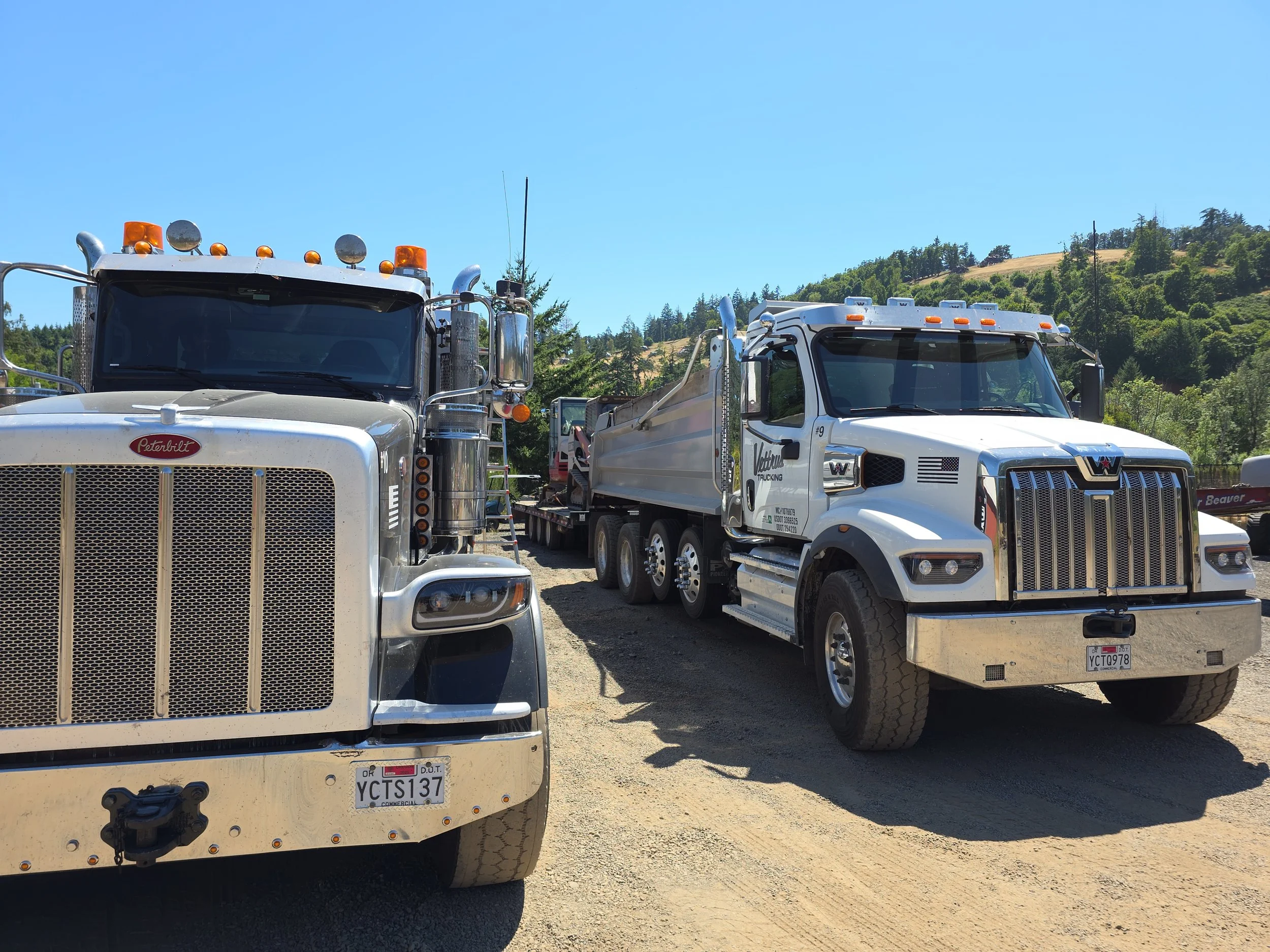 Two large trucks parked on a dirt lot against a backdrop of green hills and a blue sky.