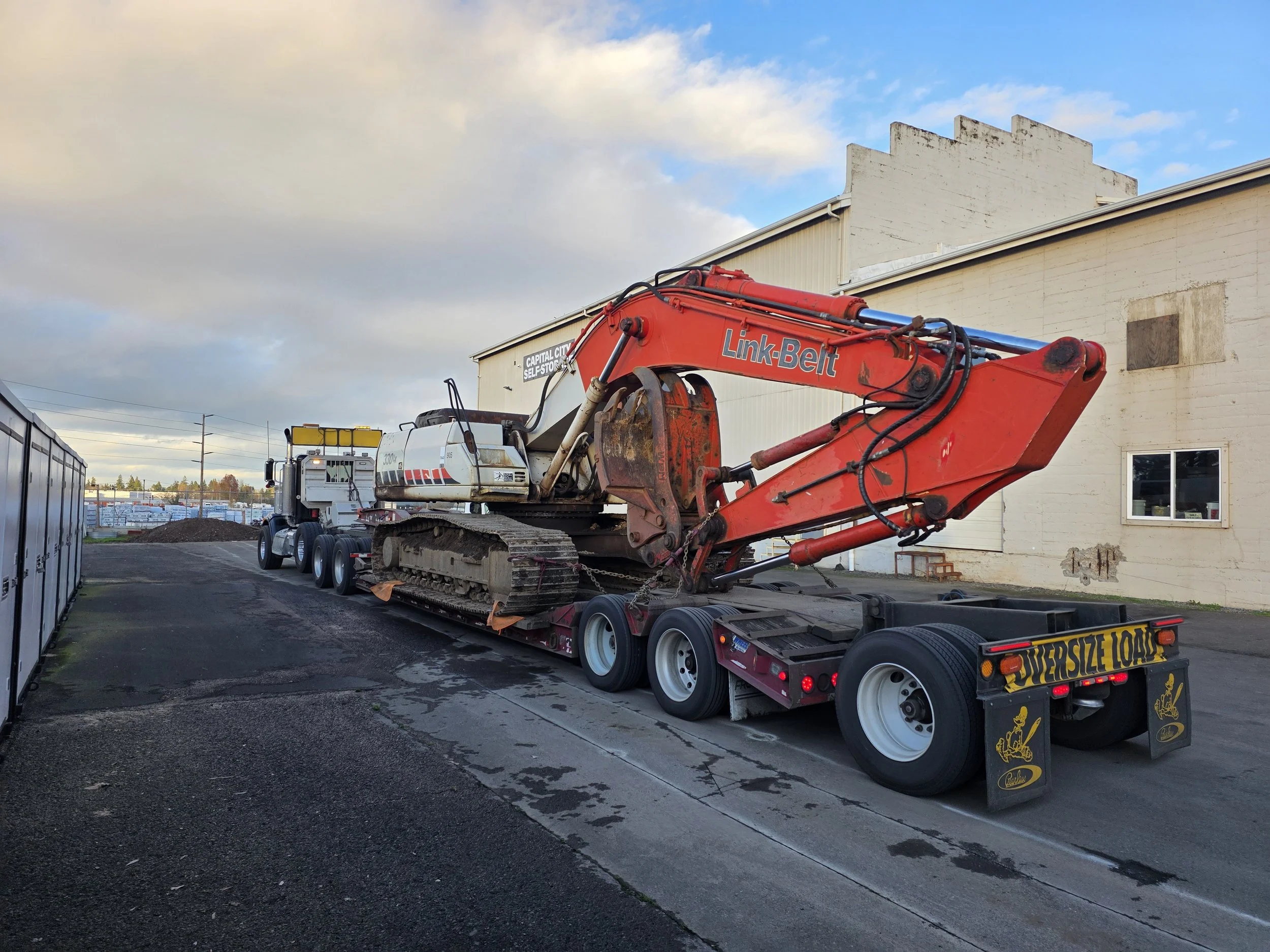 A red excavator on a flatbed truck in a parking lot with a building and cloudy sky in the background.