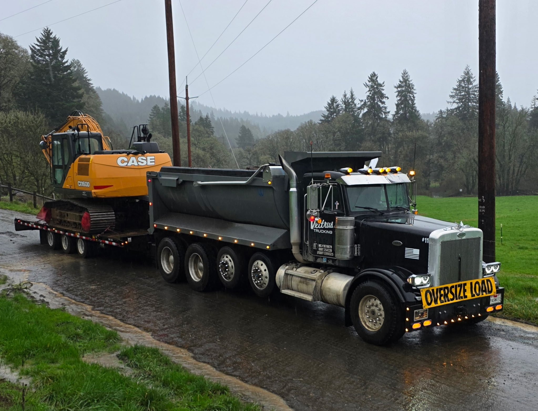 A semi-truck carrying a large yellow and black excavator on a flatbed trailer, with a rural landscape of trees and utility poles in the background, and a cloudy sky overhead. Albany Oregon.
