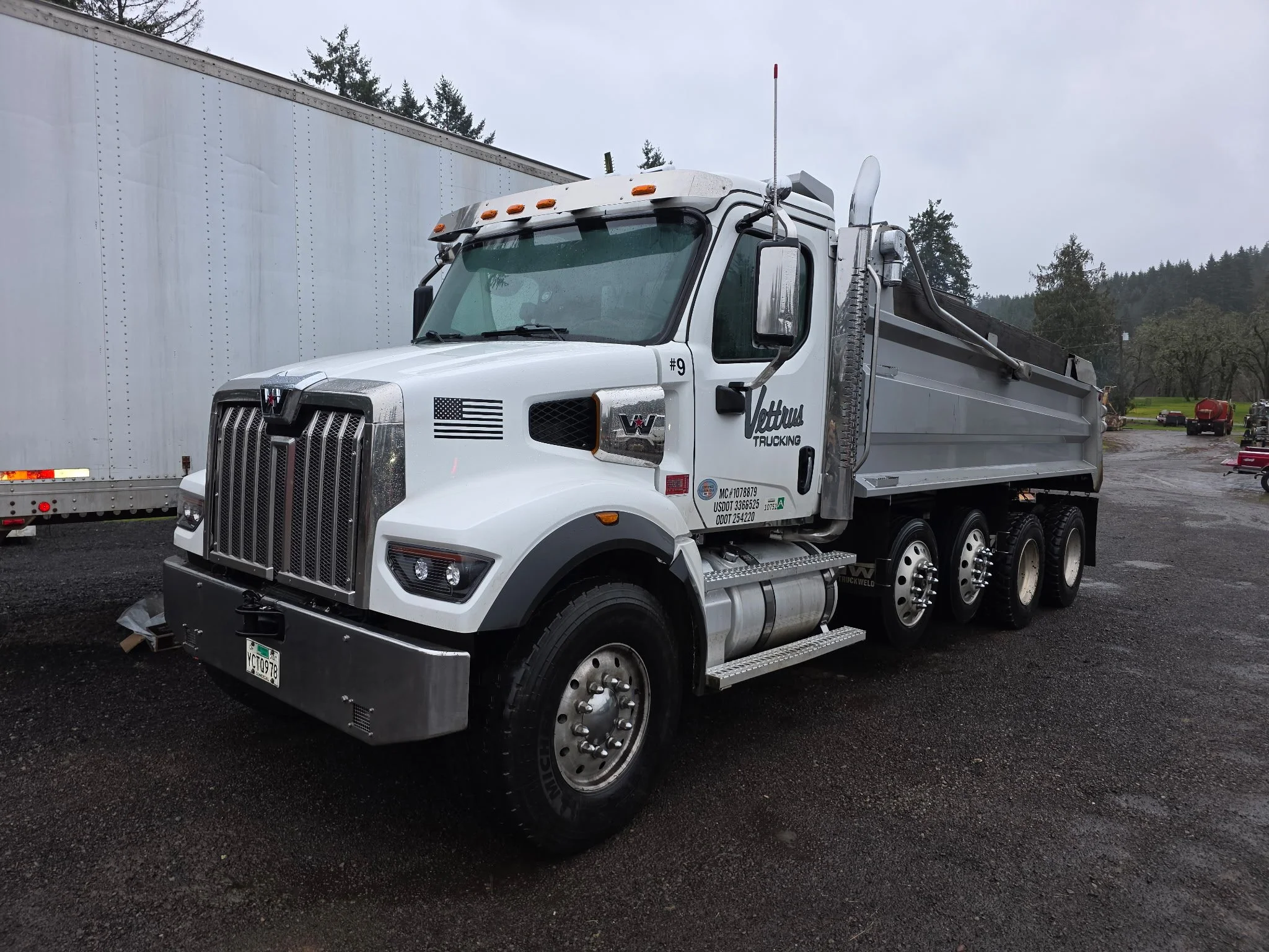A white semi-truck with a flatbed trailer parked outdoors on a cloudy day. The truck has black and white American flags decals, and the name 'Vettura Trucking' on the door. There are trees and mountains in the background.