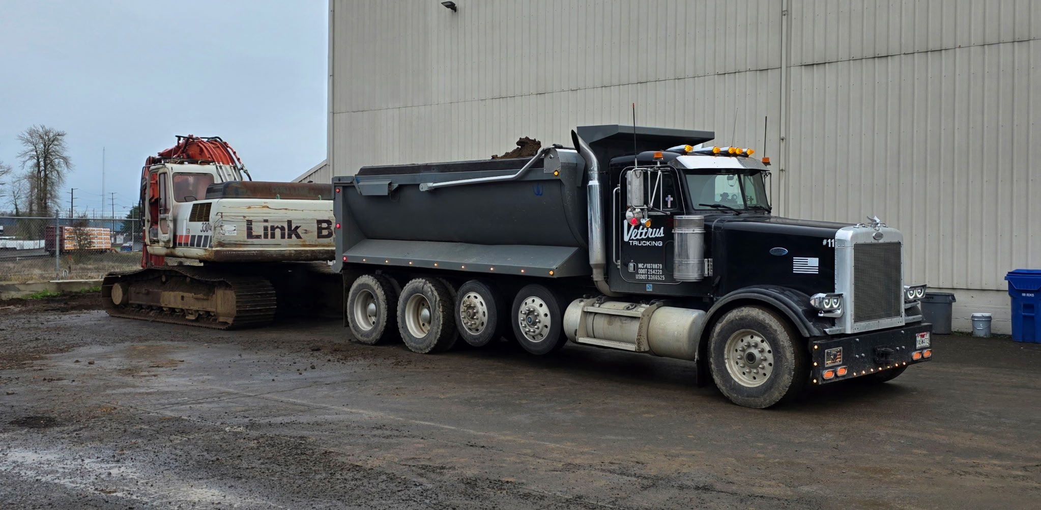 A large black and silver semi-truck with the logo 'Veturus Trucking' on the door is parked next to a bulldozer on a construction site, with a beige building wall behind them.