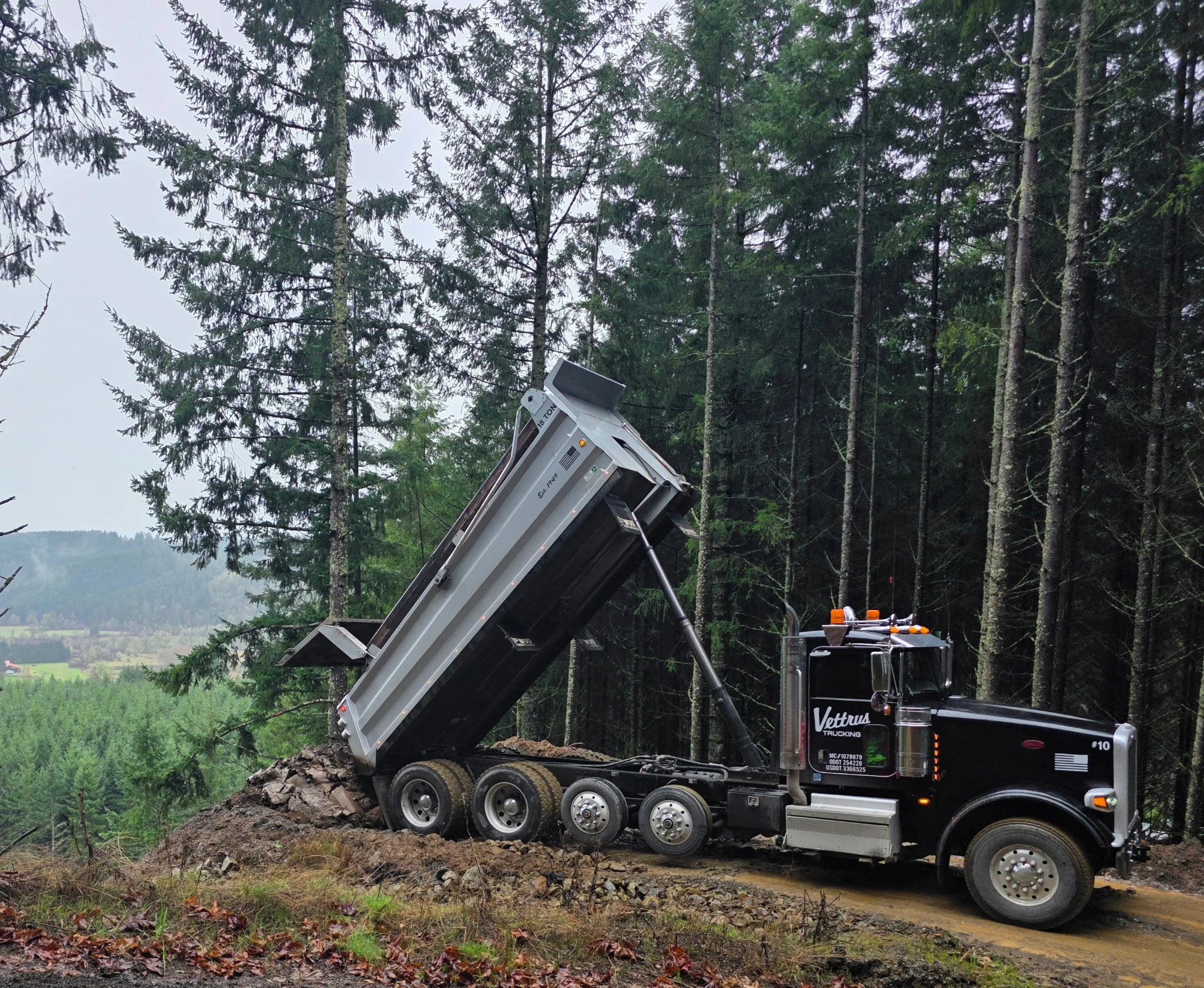 A large logging truck dumping dirt or gravel on a forested hillside.