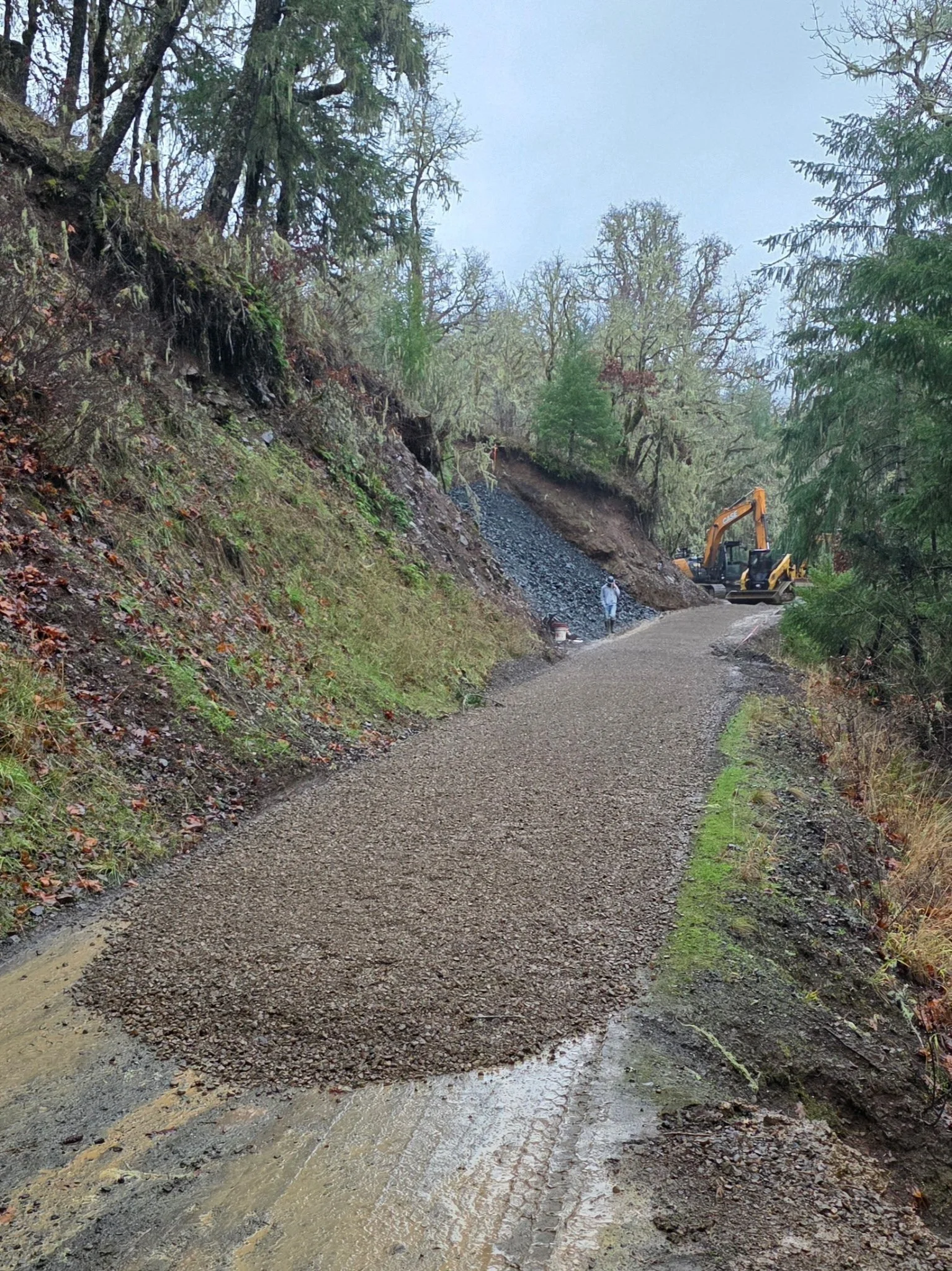 Construction workers and a small excavator next to a partially excavated hillside along a dirt road in a forested area.