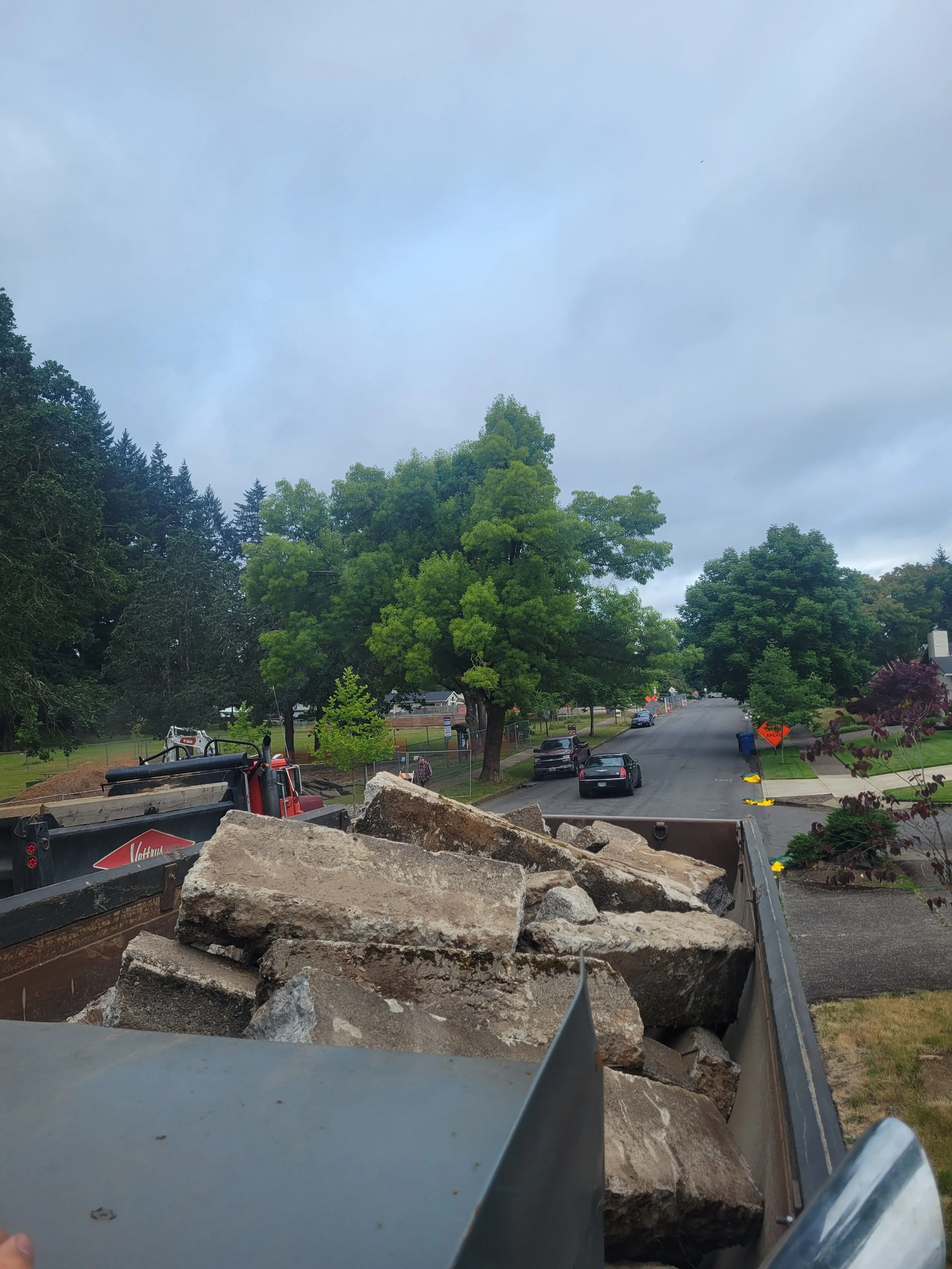 View from the back of a dump truck filled with large rocks, overlooking a residential street with parked cars, lush green trees, overcast sky, and construction signs. Salem Oregon.