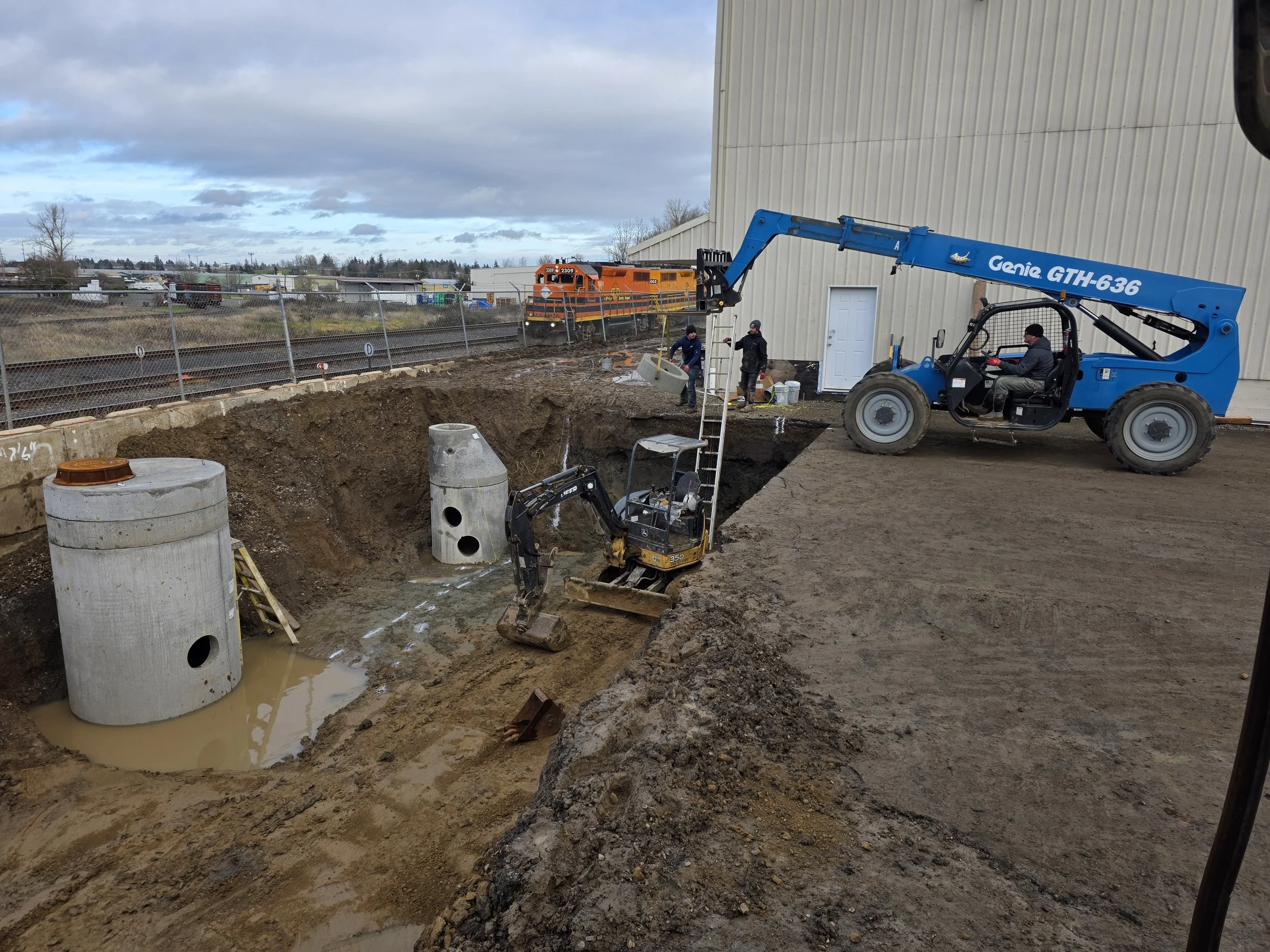 Construction site with workers near underground utility work, heavy machinery, and a train in the background under cloudy sky. Portland Oregon