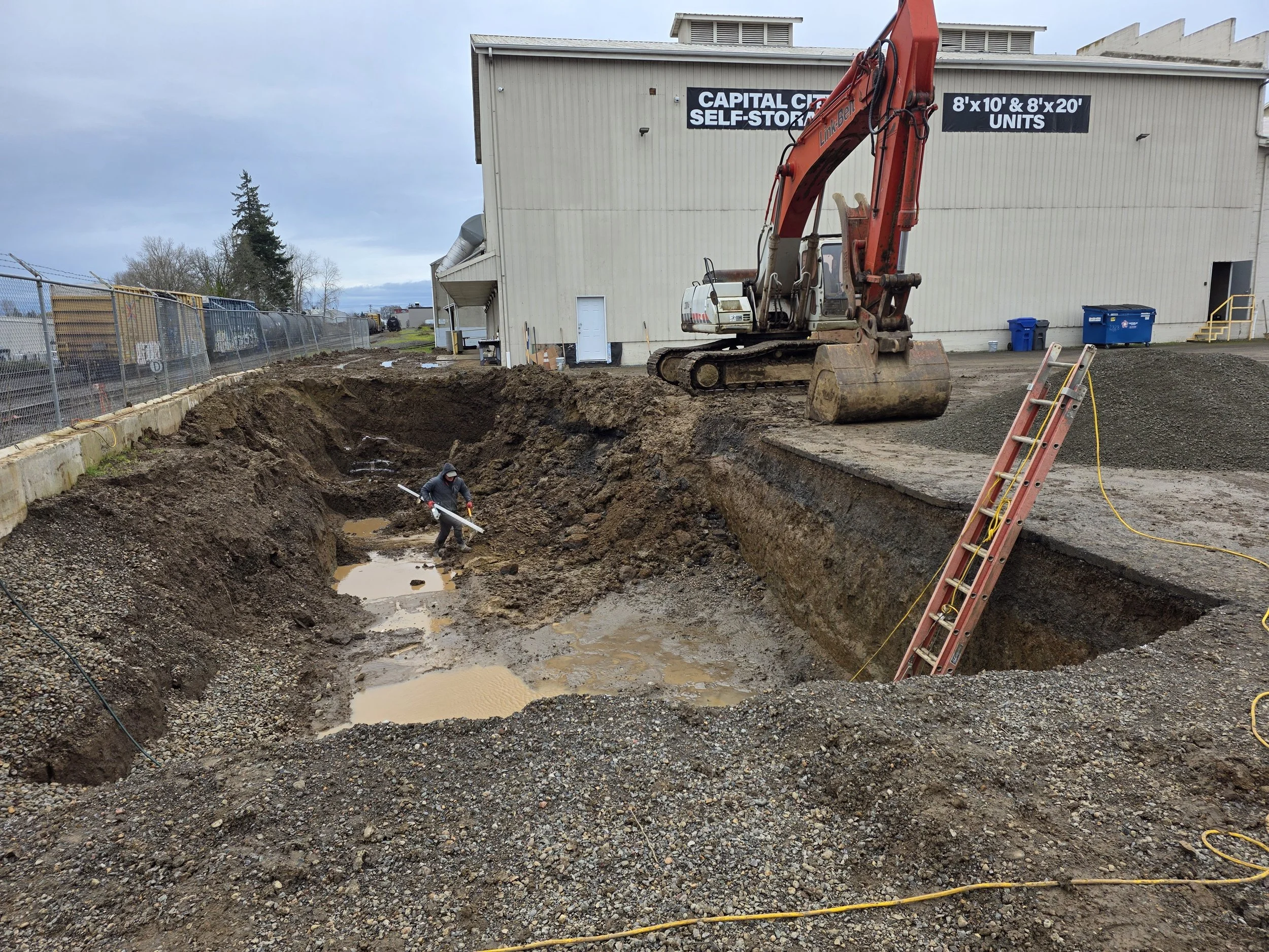 Construction site with excavator digging a trench, workers inside the trench, and a building in the background with a sign that reads 'CAPITAL CITIES SELF-STORAGE' and size options for storage units.