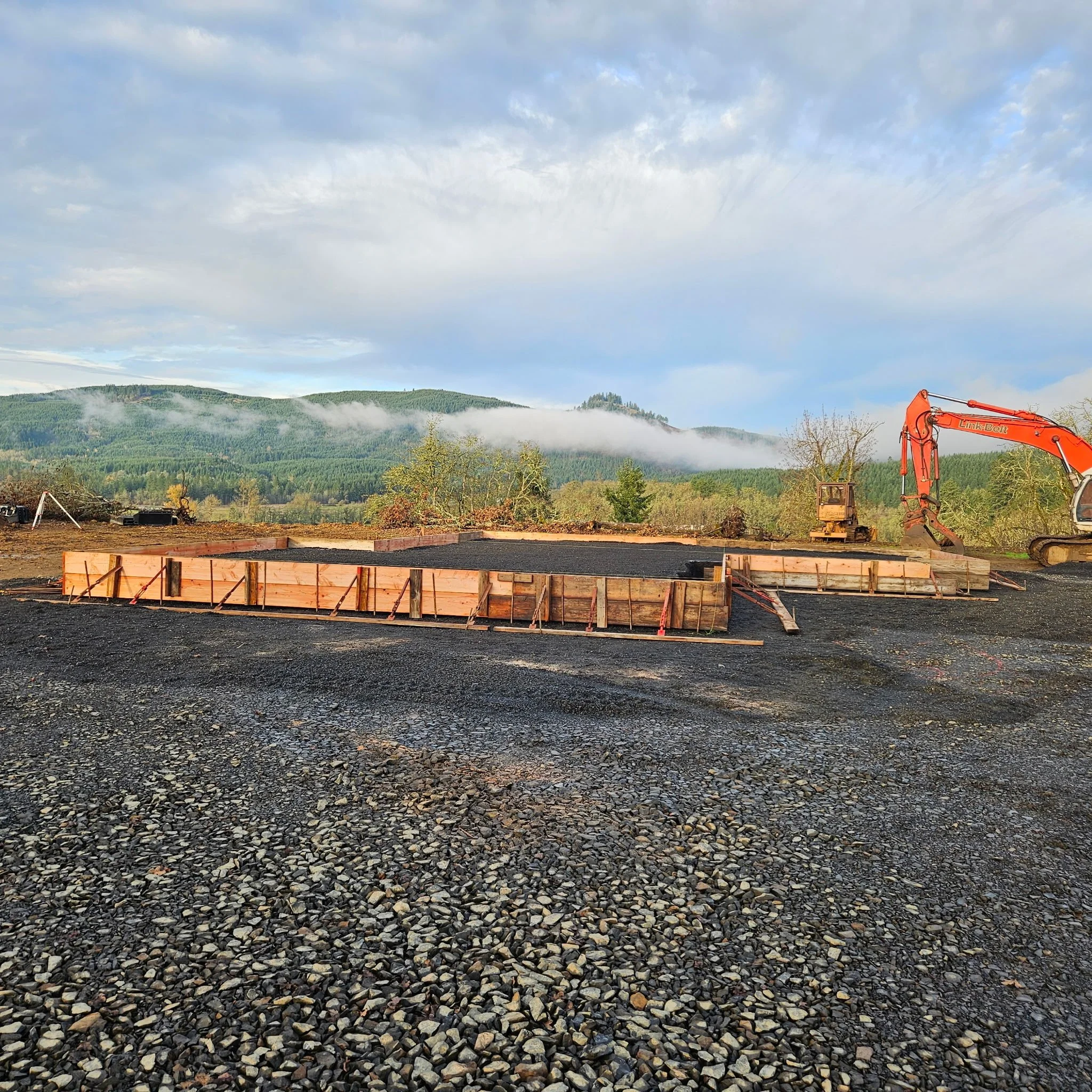 Construction site with a wooden frame foundation, an orange excavator, and a scenic background of hills and cloudy sky.