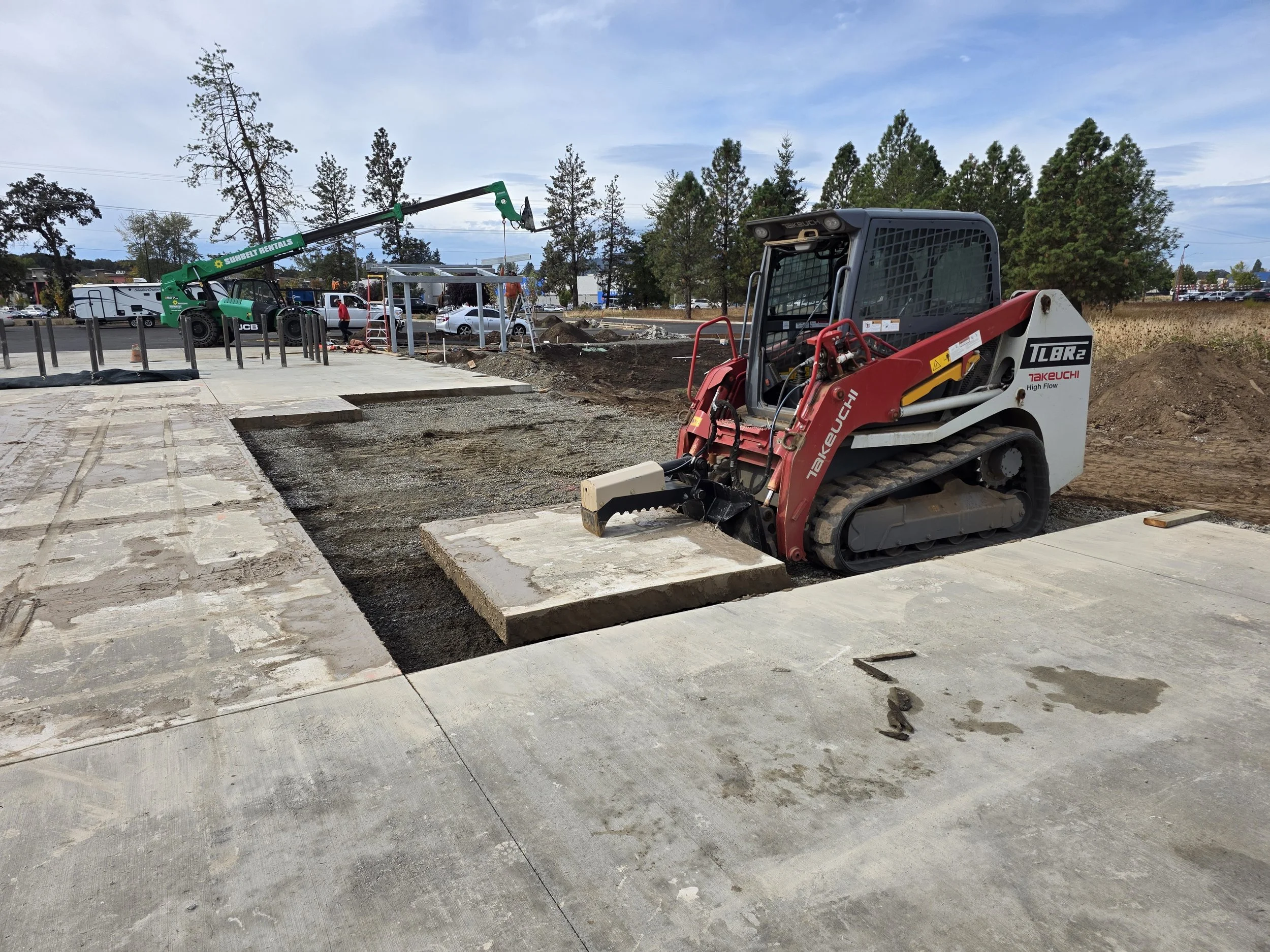 Construction site with a mini excavator and a crane lifting materials, concrete being poured, and trees in the background. Sweet Home Oregon 