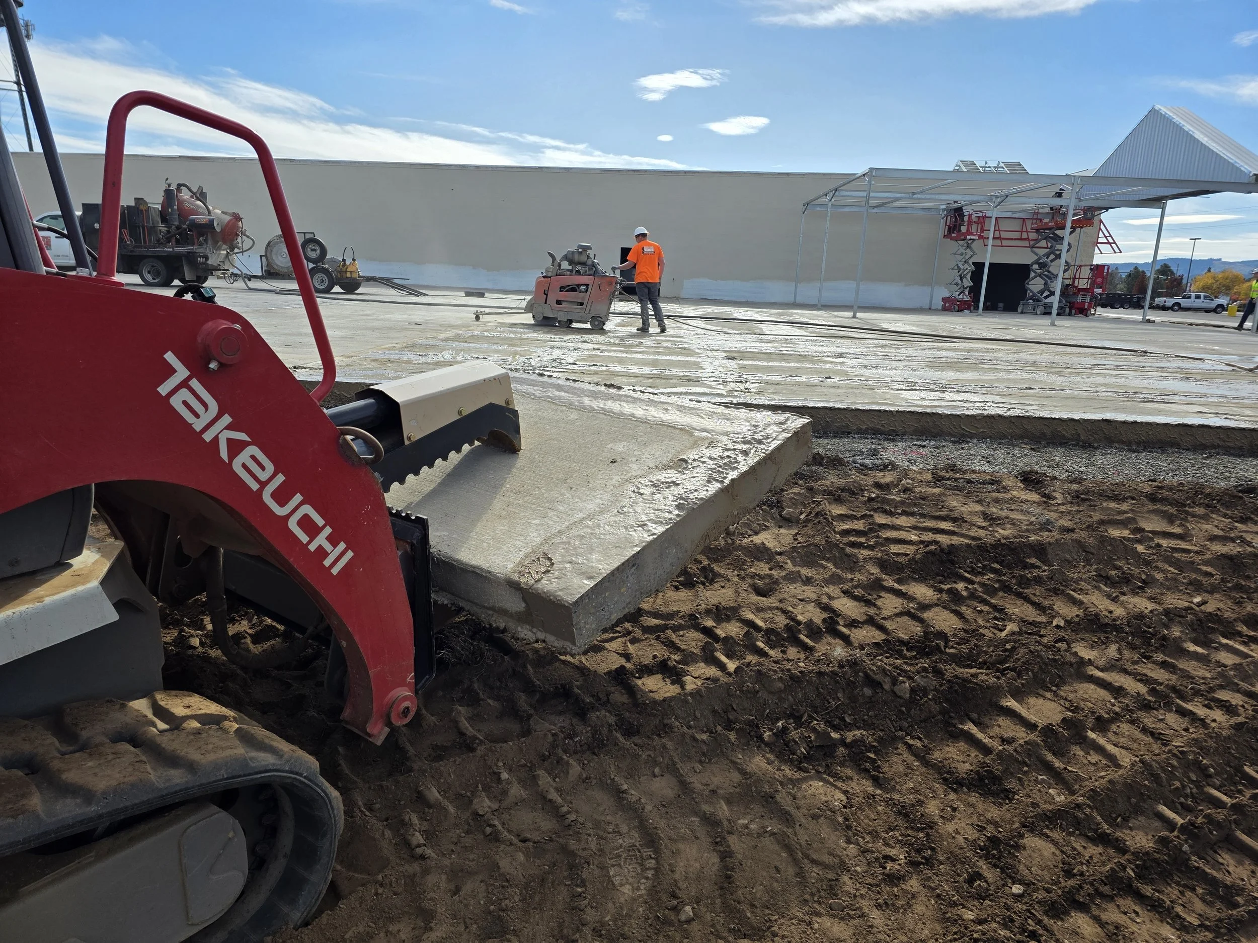 Construction worker operating a large floor grinder on a concrete surface at an outdoor building site with construction equipment and a partially built structure nearby.