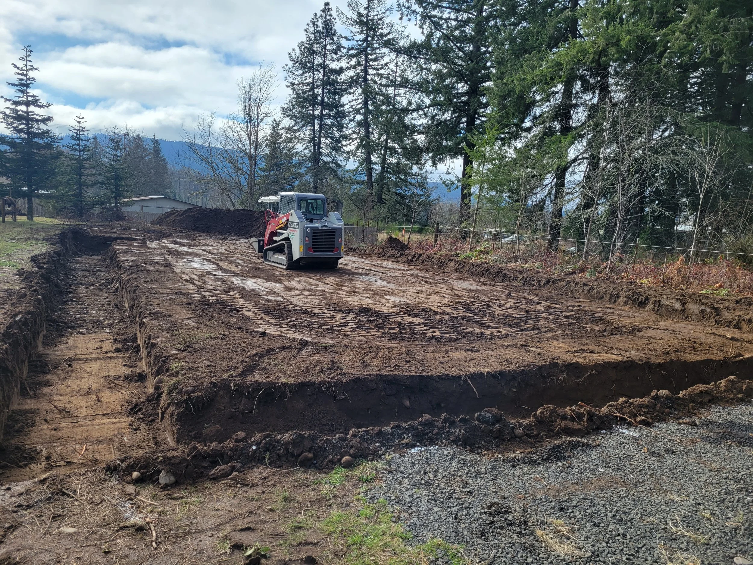 A construction site with a small excavator moving dirt, surrounded by tall trees and mountains in the background.