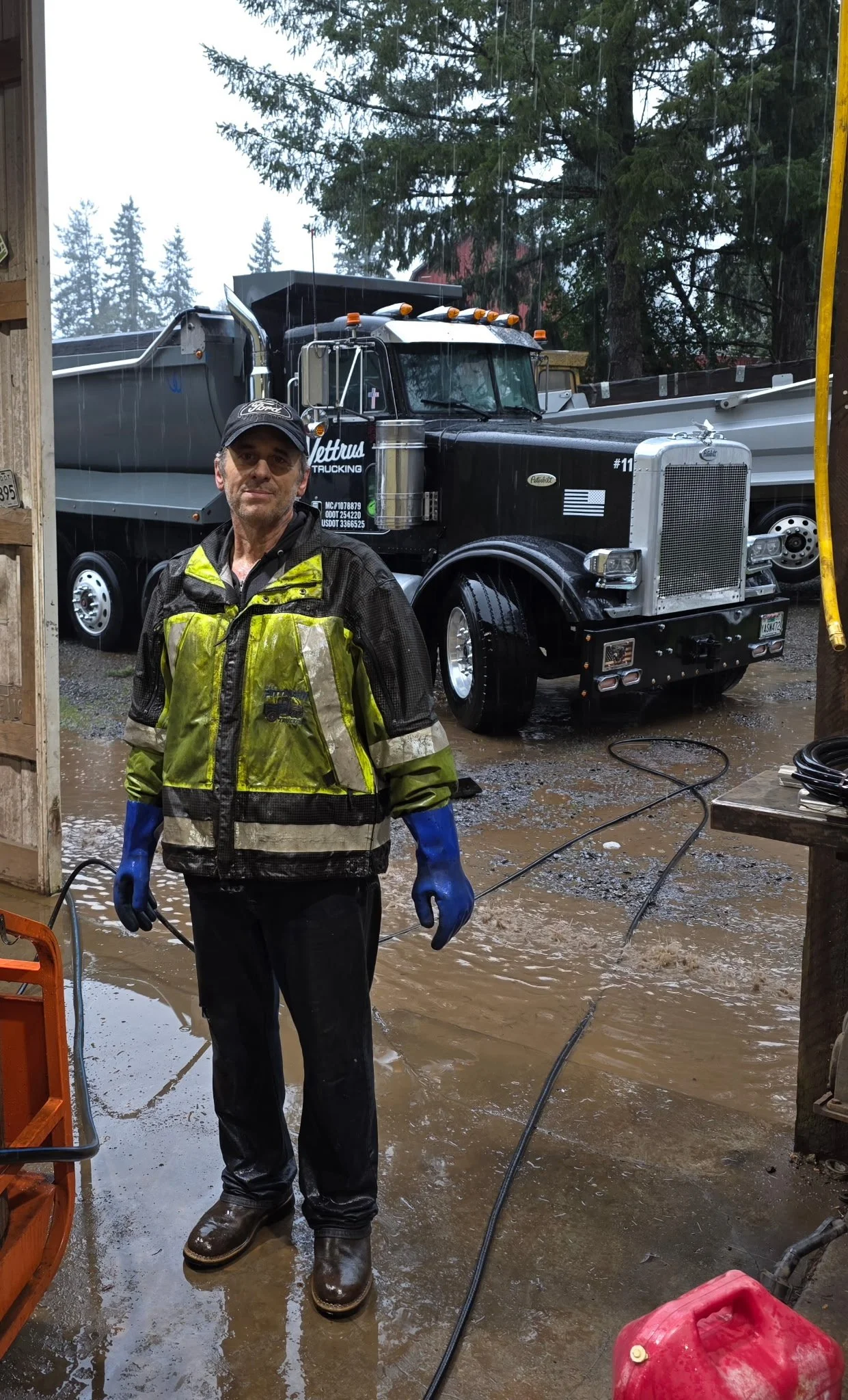 A man wearing a black Ford cap, a yellow and black waterproof jacket, and blue gloves stands in a flooded garage with muddy water on the ground, a large black and silver dump truck is parked behind him, and trees are visible in the background.