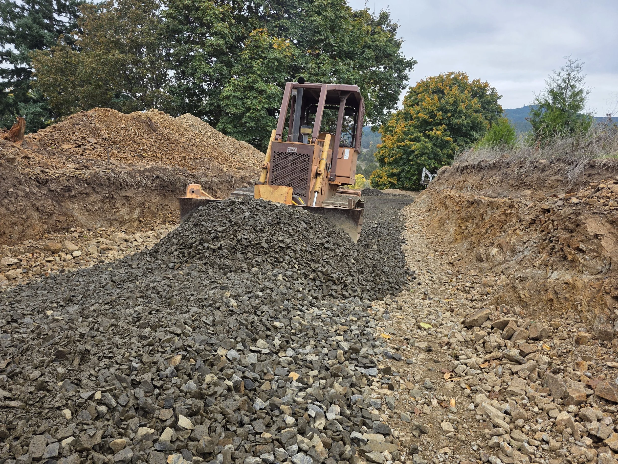 Construction site with dirt and gravel, a small bulldozer, and trees in the background.
