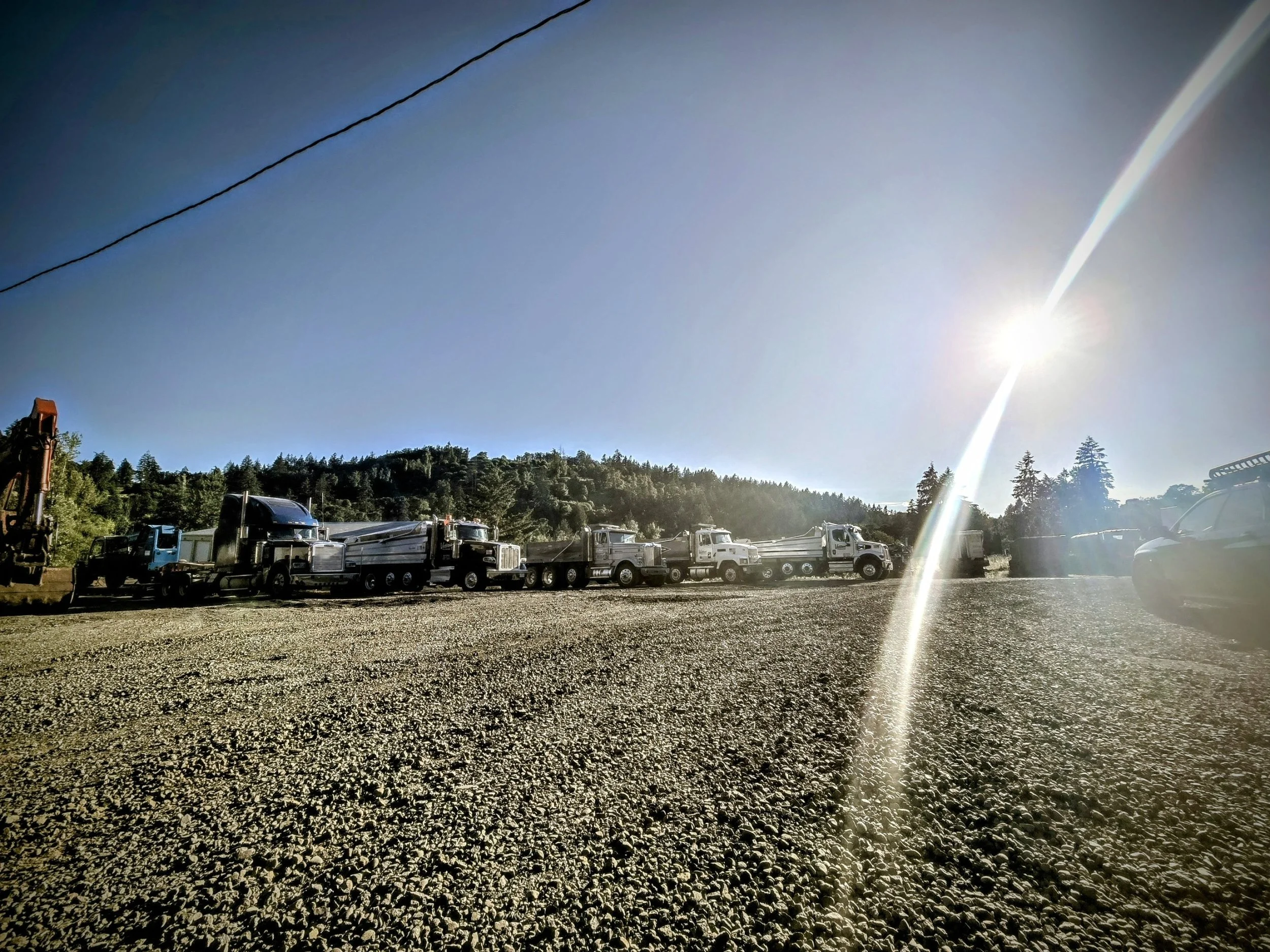 Line of trucks parked outdoors under a bright sun with clear blue sky, trees, and hills in the background.