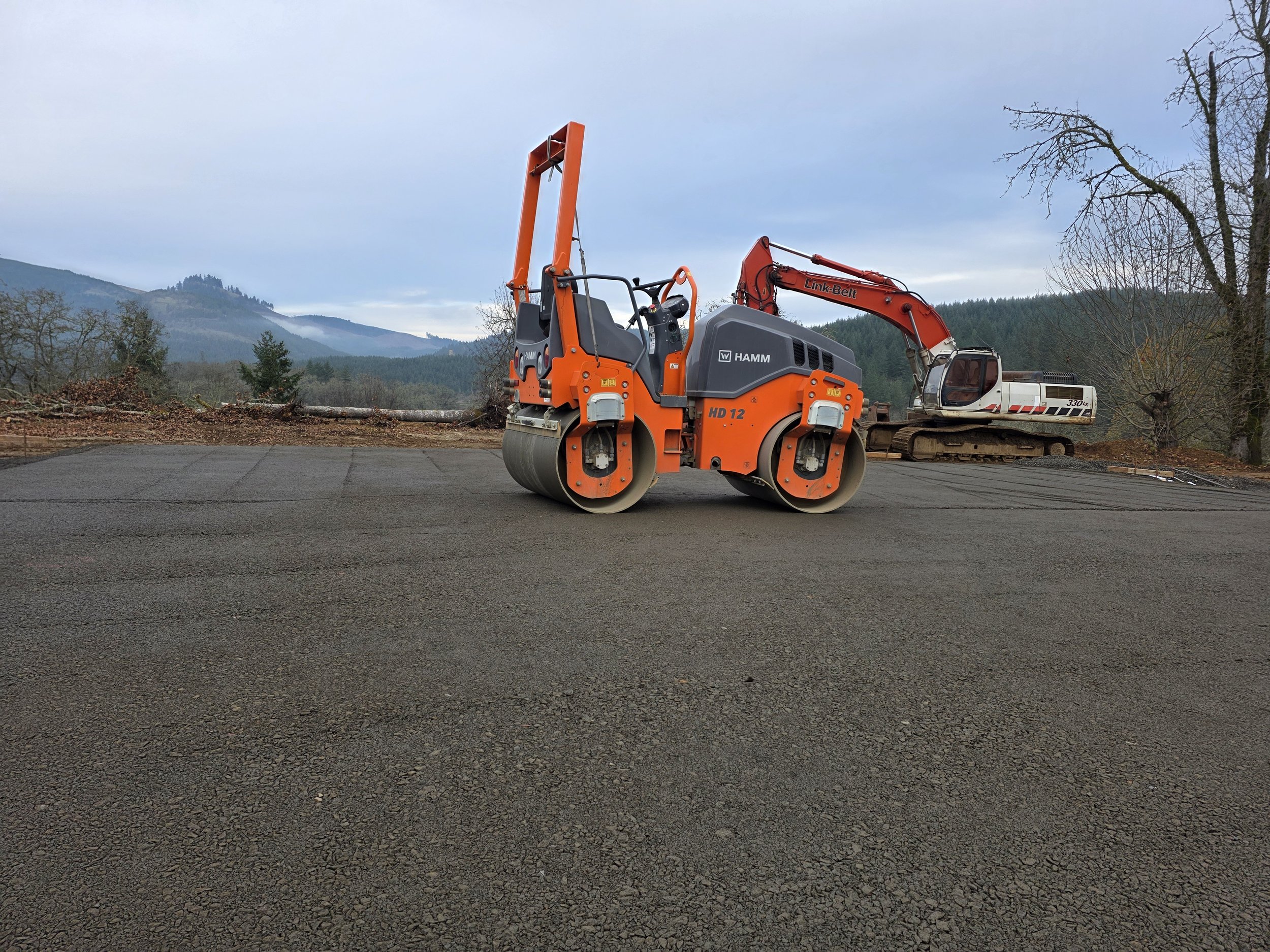 Construction scene with heavy machinery, including a large orange roller compactor and a white excavator, set against a backdrop of distant mountains and cloudy sky.
