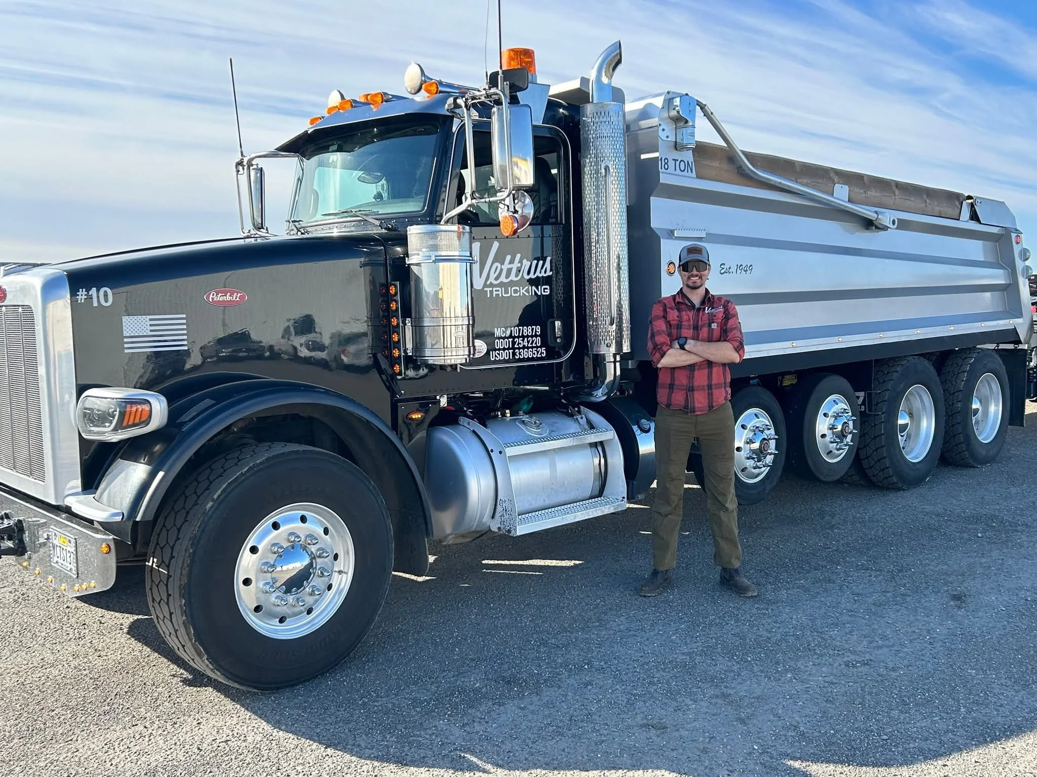 A man in sunglasses and a red plaid shirt standing next to a large black and silver dump truck with the Vettus Trucking logo, American flag, and various identification numbers on the side, in an open outdoor area with a blue sky and few clouds.