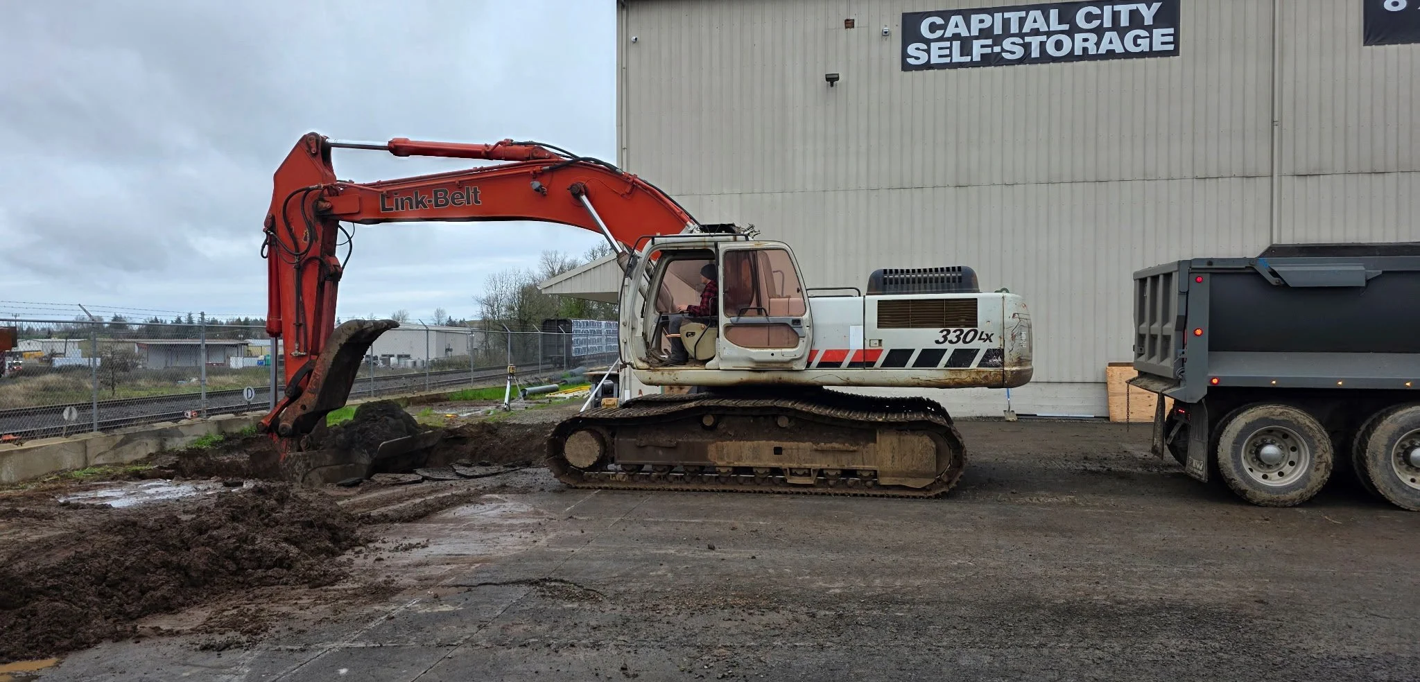 A construction excavator digging at a site in front of a building with a sign that reads 'Capital City Self-Storage'.
