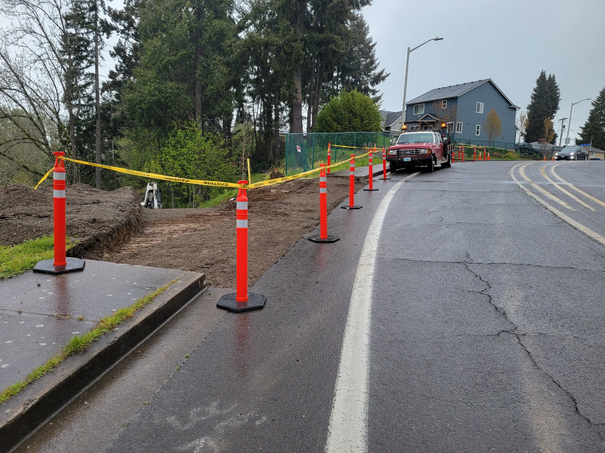 A street with road work in progress. Orange cones and yellow caution tape block part of the sidewalk and road. A red utility truck is parked on the street next to the construction area. Green trees and a house are visible in the background on a cloud