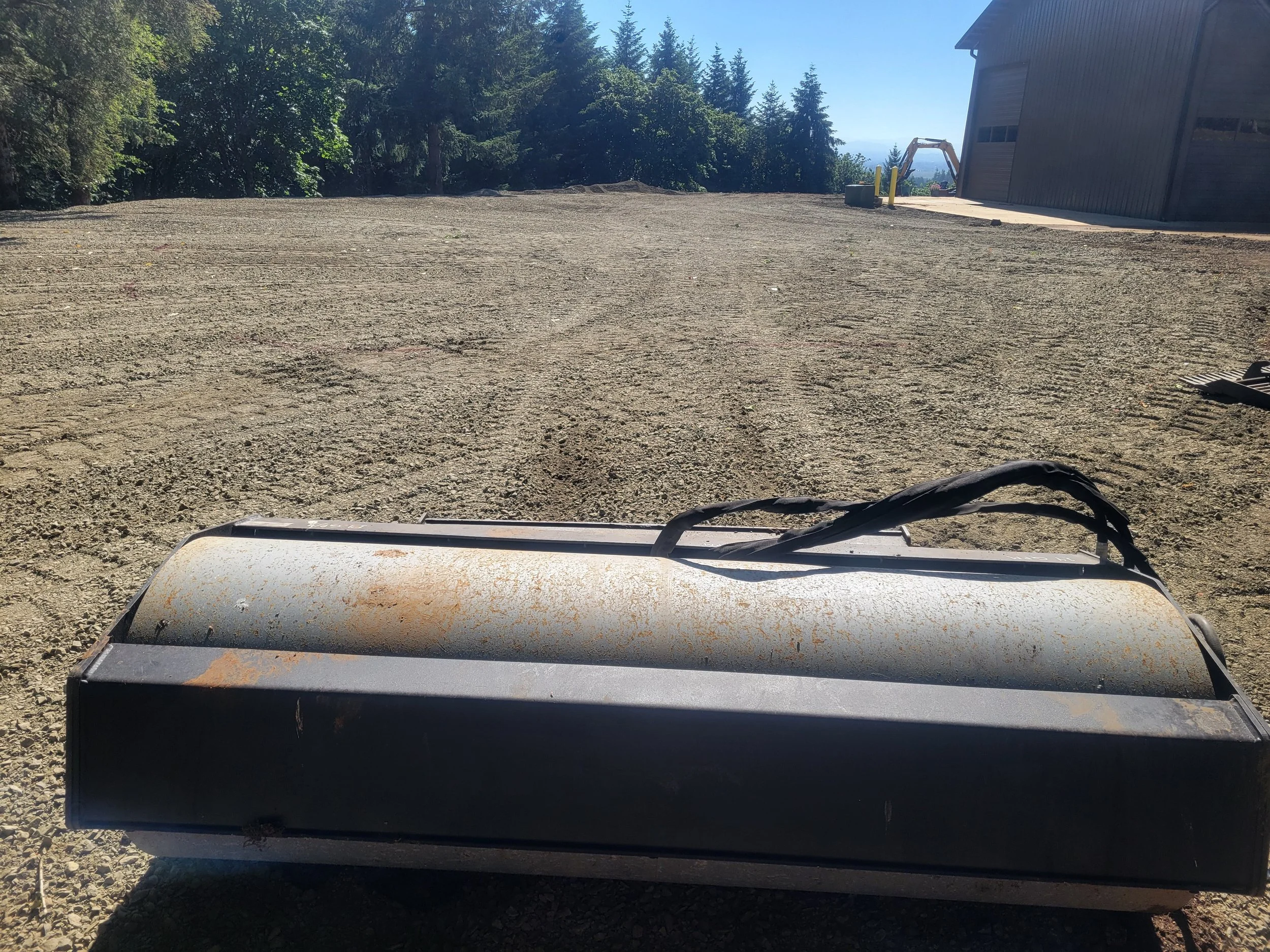 A cleared dirt lot with a background of trees, a building on the right, and a construction excavator in the distance. Part of a machine or equipment is visible in the foreground. Salem Oregon