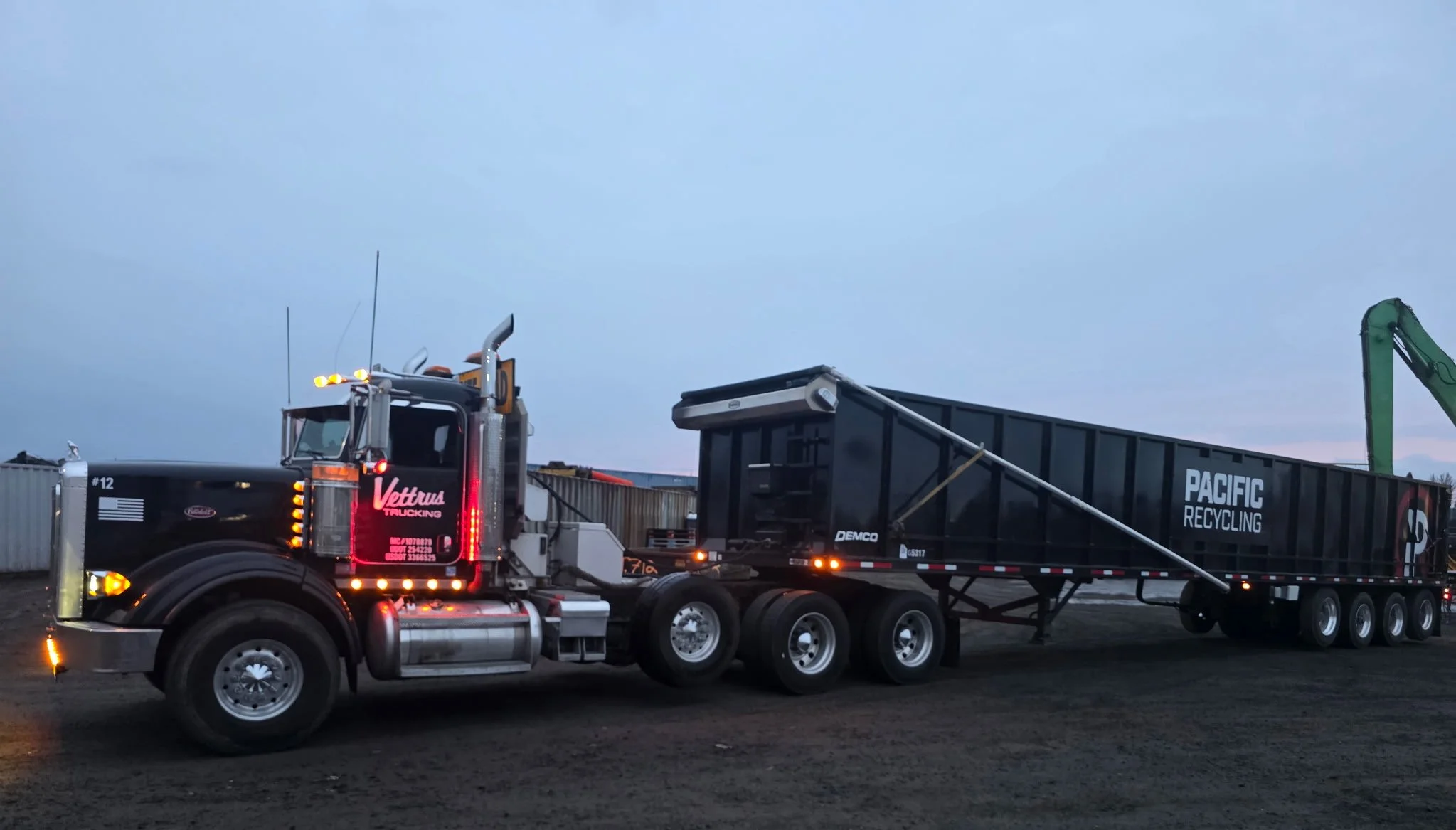 A black semi-truck with orange and white accents, labeled Vettrus Trucking, hauling a large black recycling container from Pacific Recycling, with a green excavator arm visible on the right side, set against a cloudy sky at dusk. Salem Oregon