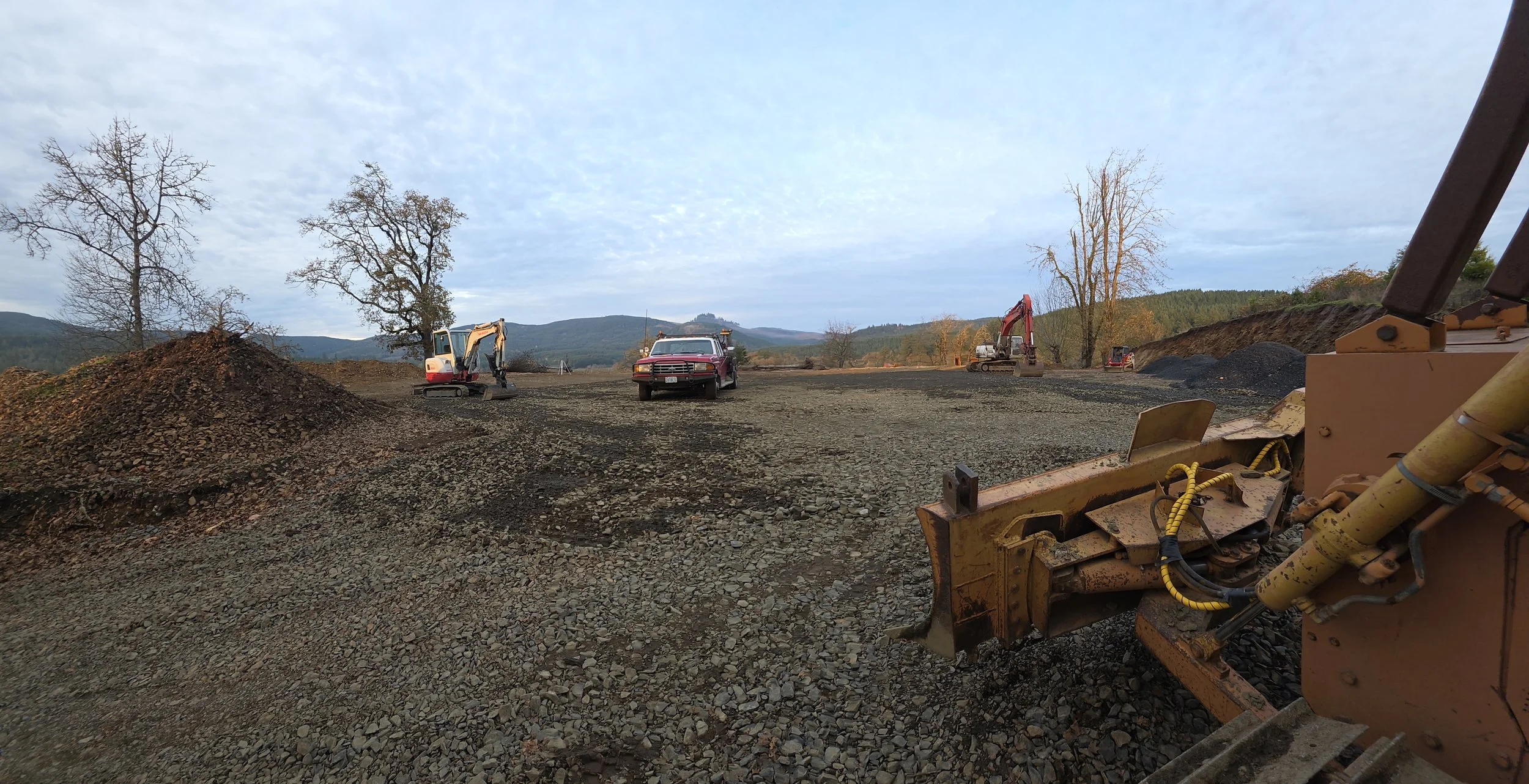Construction site with dirt piles, two mini excavators, a red pickup truck, and large machinery, set against a backdrop of trees and distant hills under a cloudy sky.