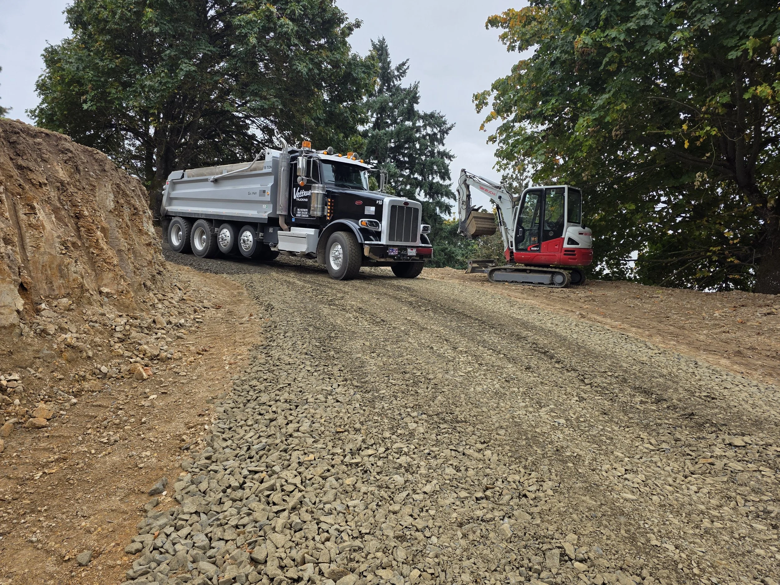 A black dump truck and a small red and white excavator working on a dirt and gravel construction site surrounded by green trees.