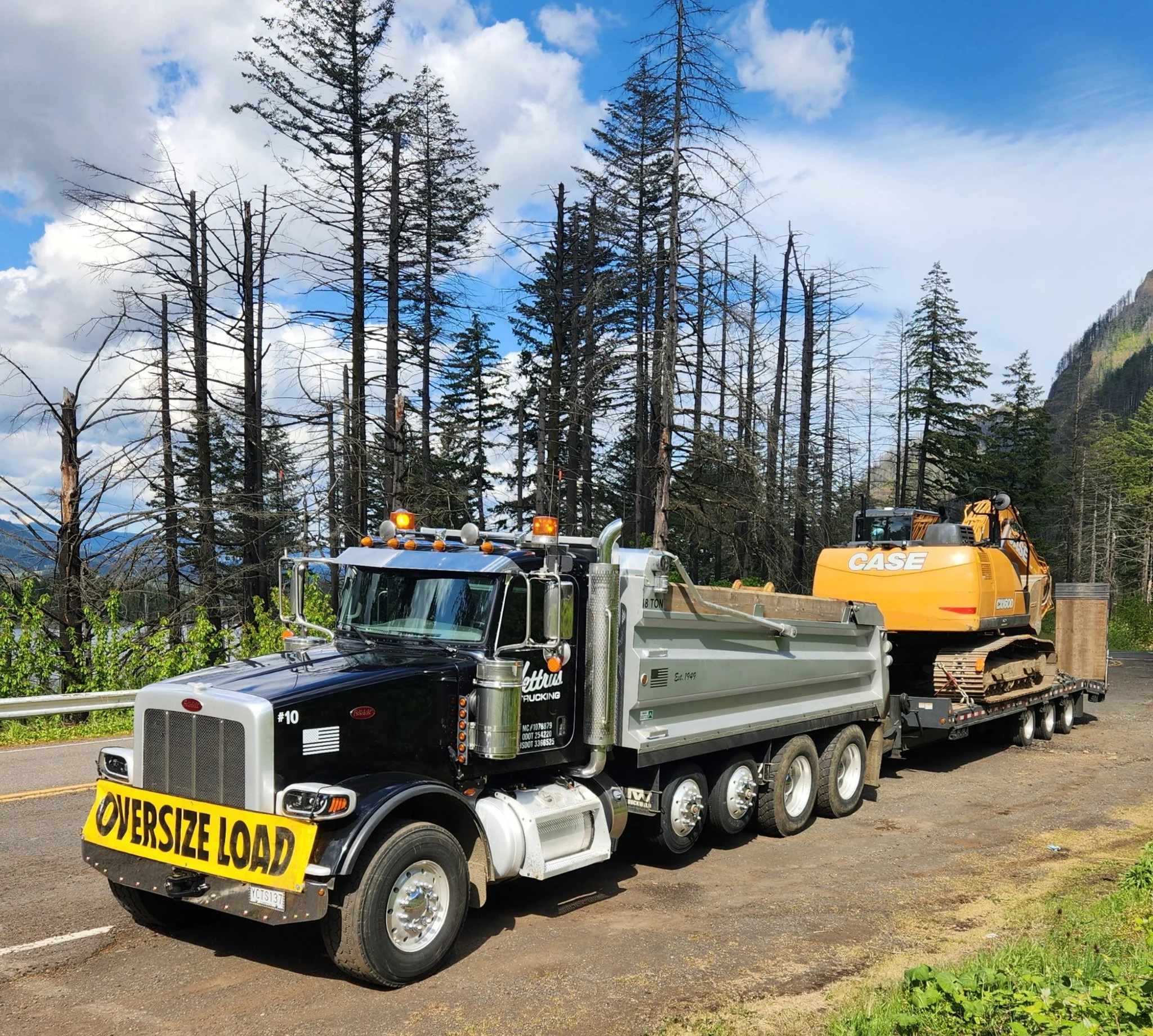 Construction truck with a yellow sign that reads 'Over Sized Load' parked on a forested roadside, with a backhoe loader visible in the background, tall trees, and a partly cloudy sky.