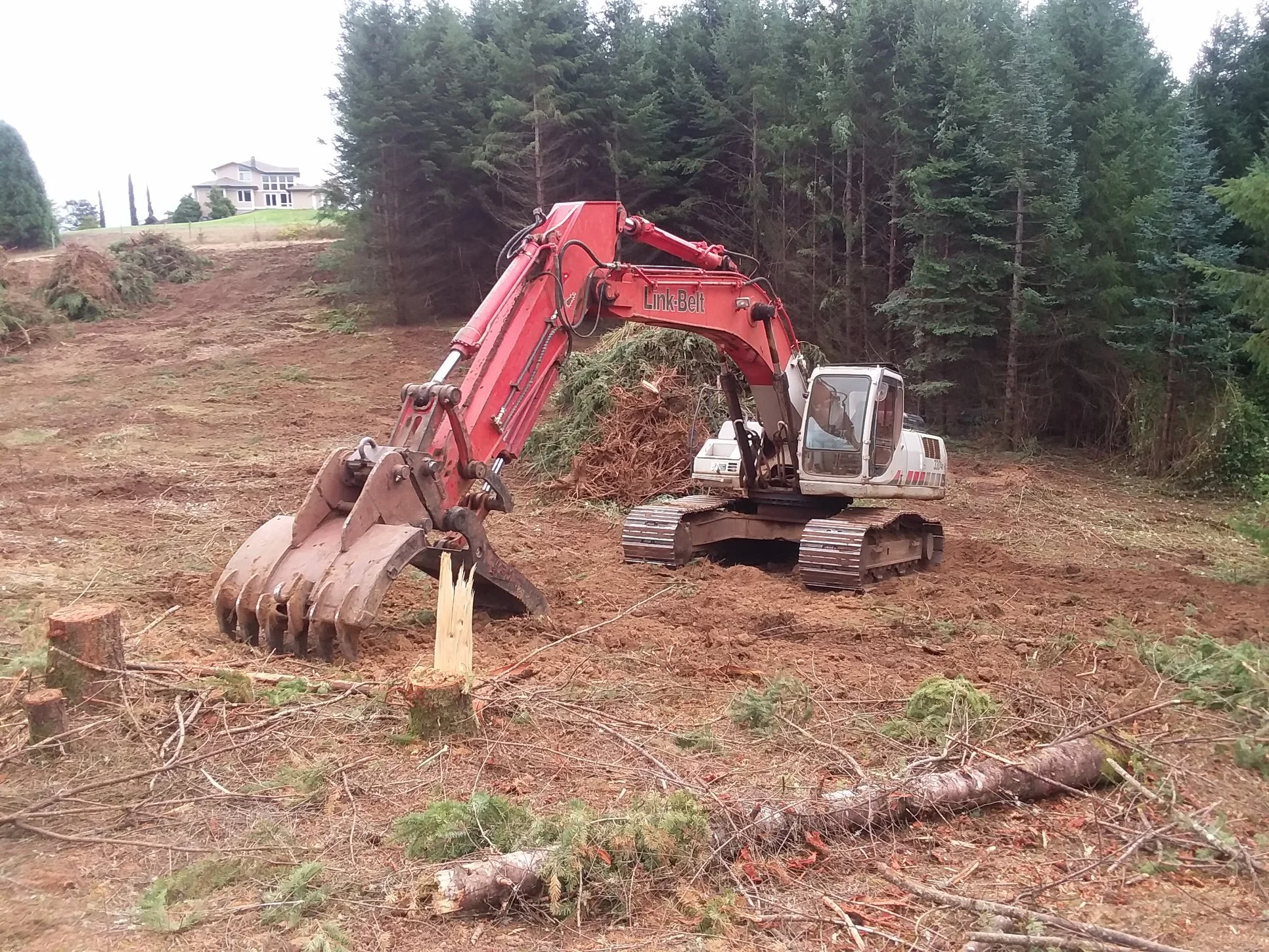 A construction excavator clearing trees on a hillside near a residential area with a house in the background. Jefferson Oregon.