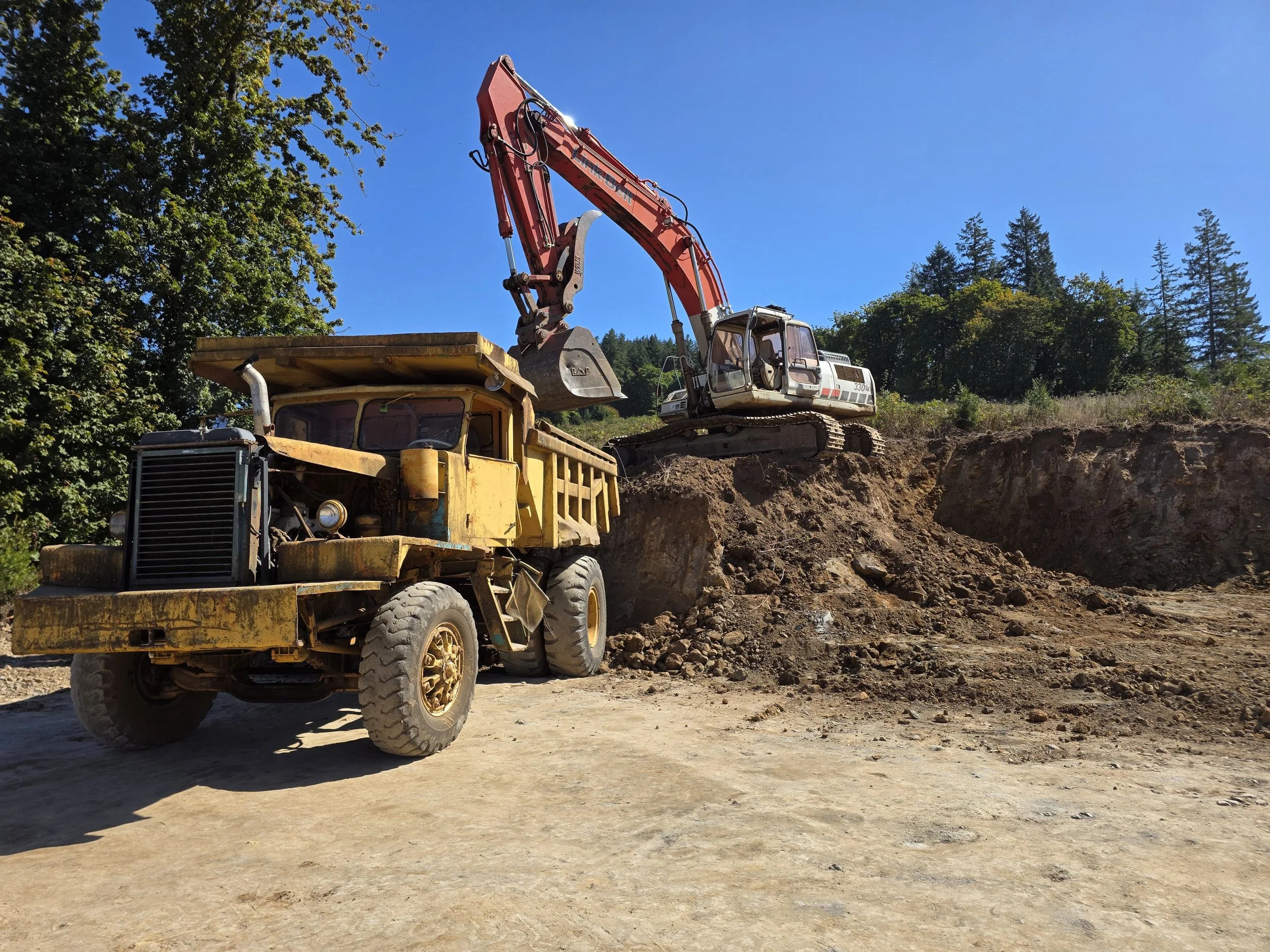 A large orange excavator with a bucket attachment dumping dirt into the bed of a yellow dump truck on a construction site surrounded by trees.
