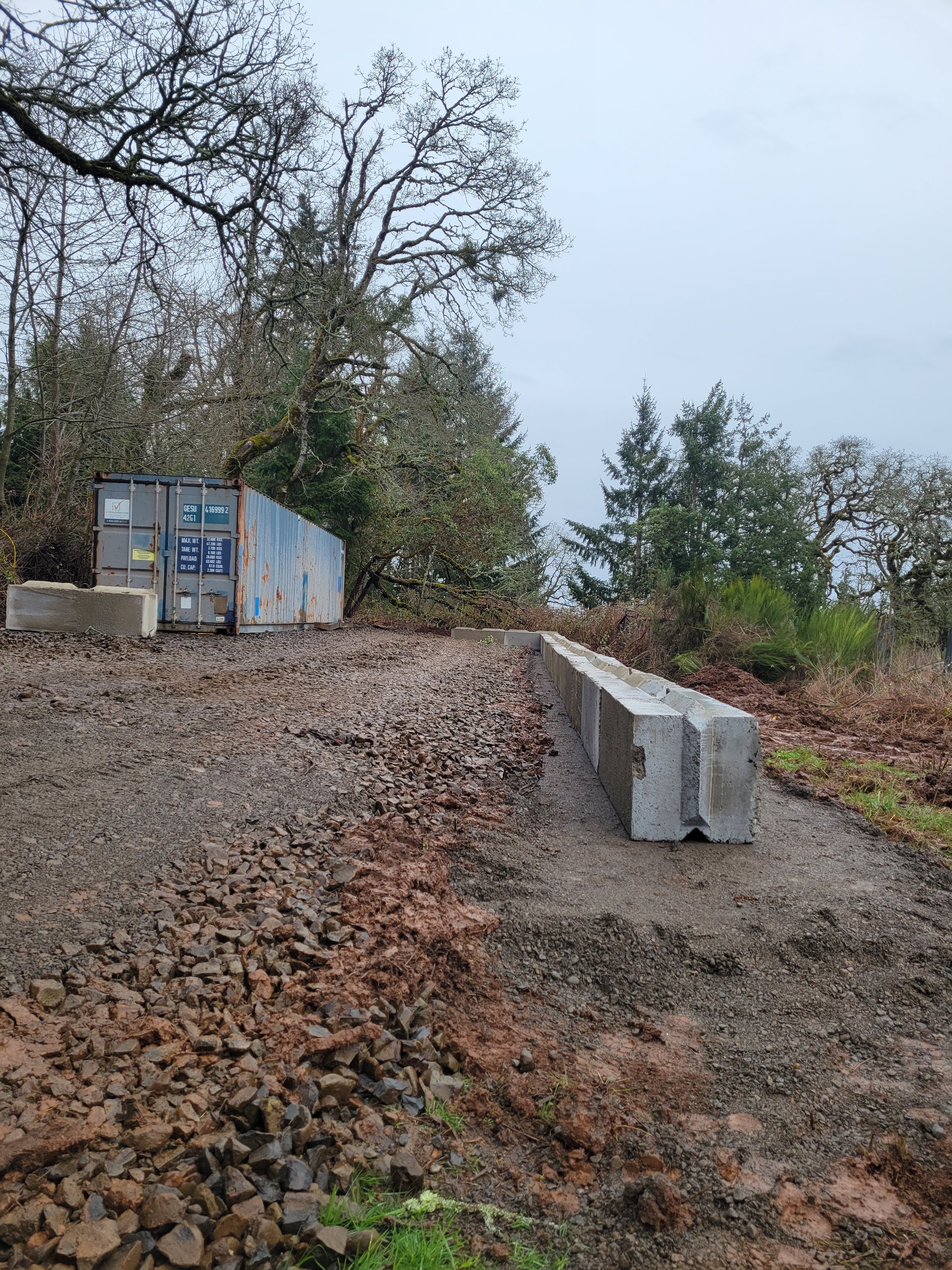 A construction site with a gravel road, a storm drain block, and a concrete barrier on the side. There are trees with bare branches and some evergreen trees in the background, with an overcast sky.