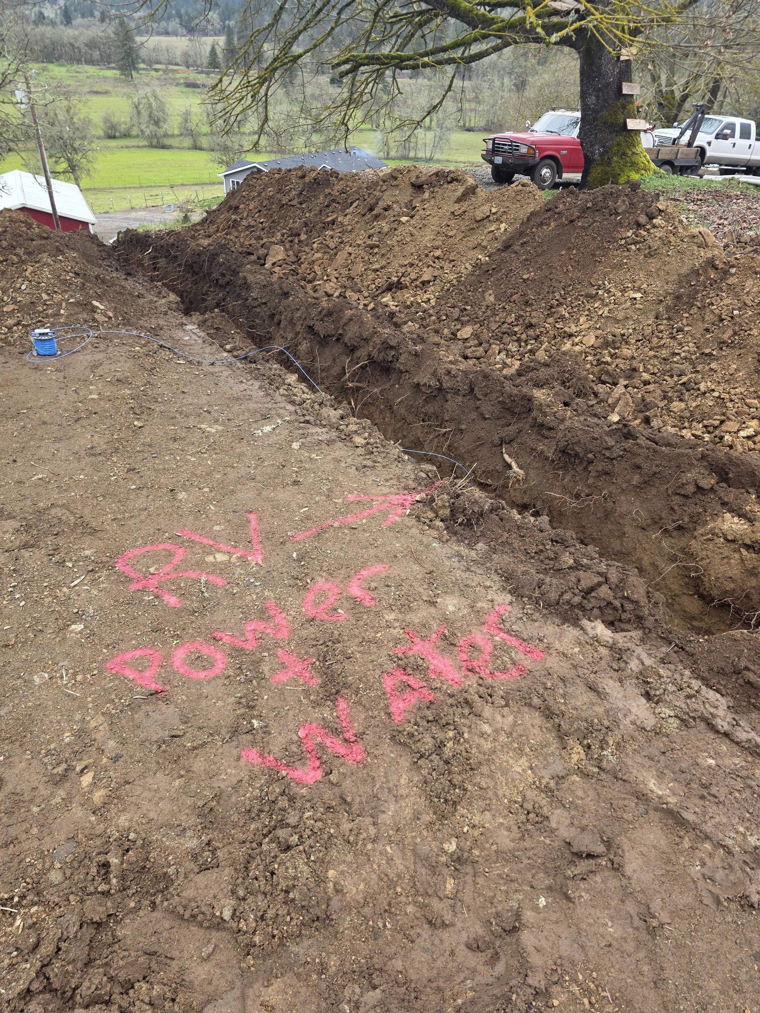 A construction site in a rural area with a large trench dug in the dirt. Pink spray-painted words are visible on the ground, saying 'POW!' and possibly other words. A blue spool of wire or cable is on the ground near the trench. In the background, th