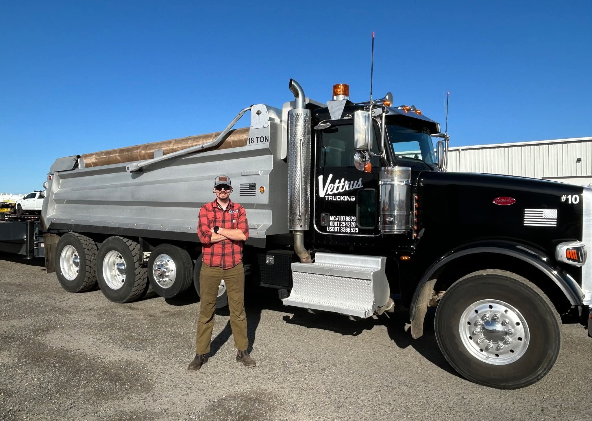 A man in a red plaid shirt, khaki pants, and a white cap with sunglasses, standing with arms crossed in front of a large black and silver dump truck with multiple wheels and orange lights on the cabin, in an outdoor industrial area under a clear blue
