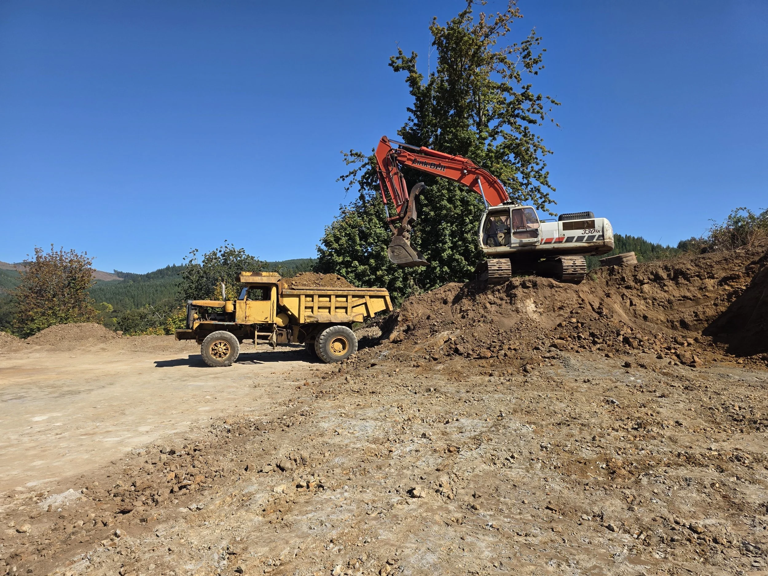 Construction site with a large excavator and a dump truck on uneven dirt terrain, with trees and hills in the background.