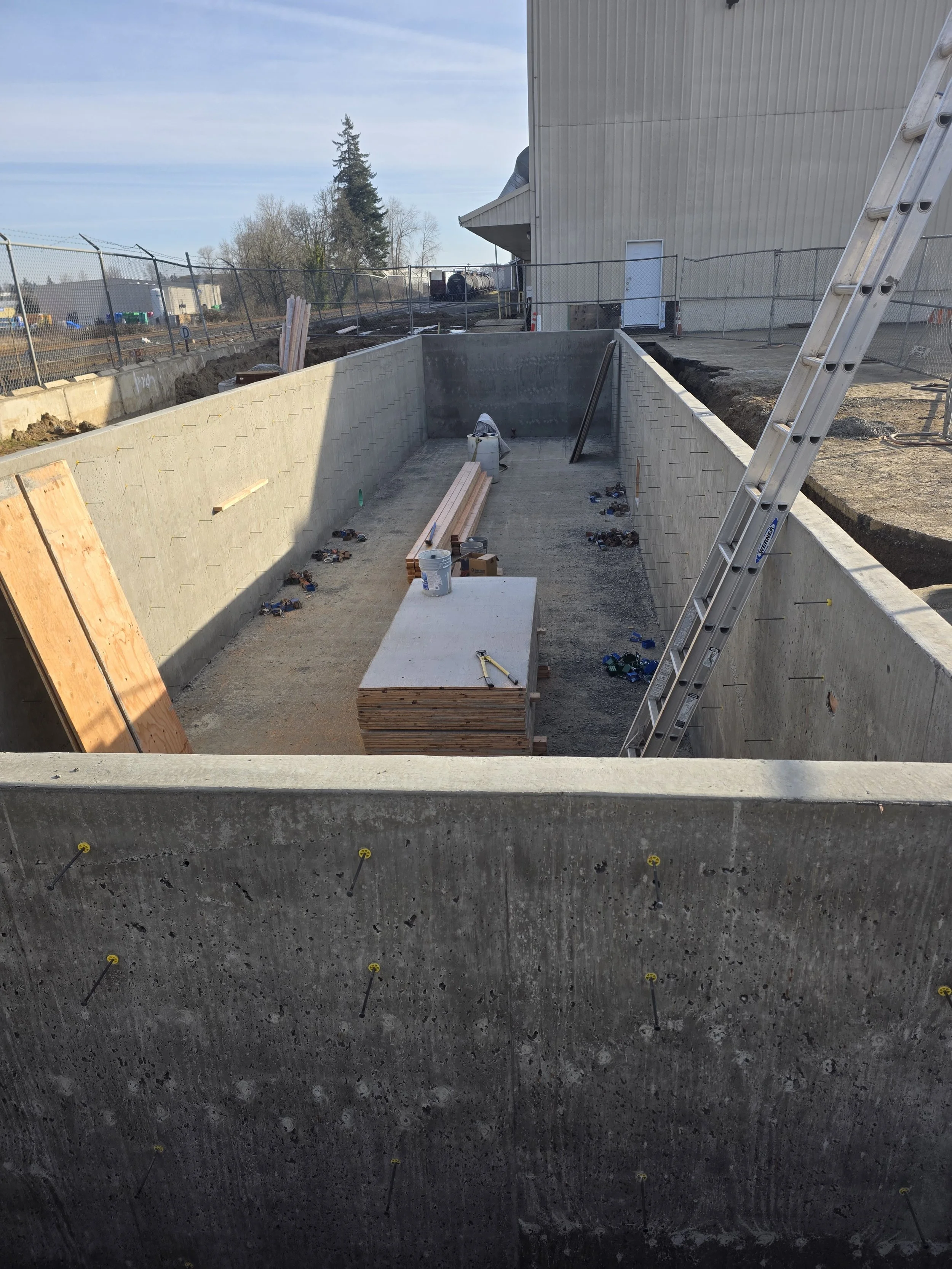 Construction site with concrete pool outline, leaning ladder, wooden planks, and tools, next to a beige building under construction.
