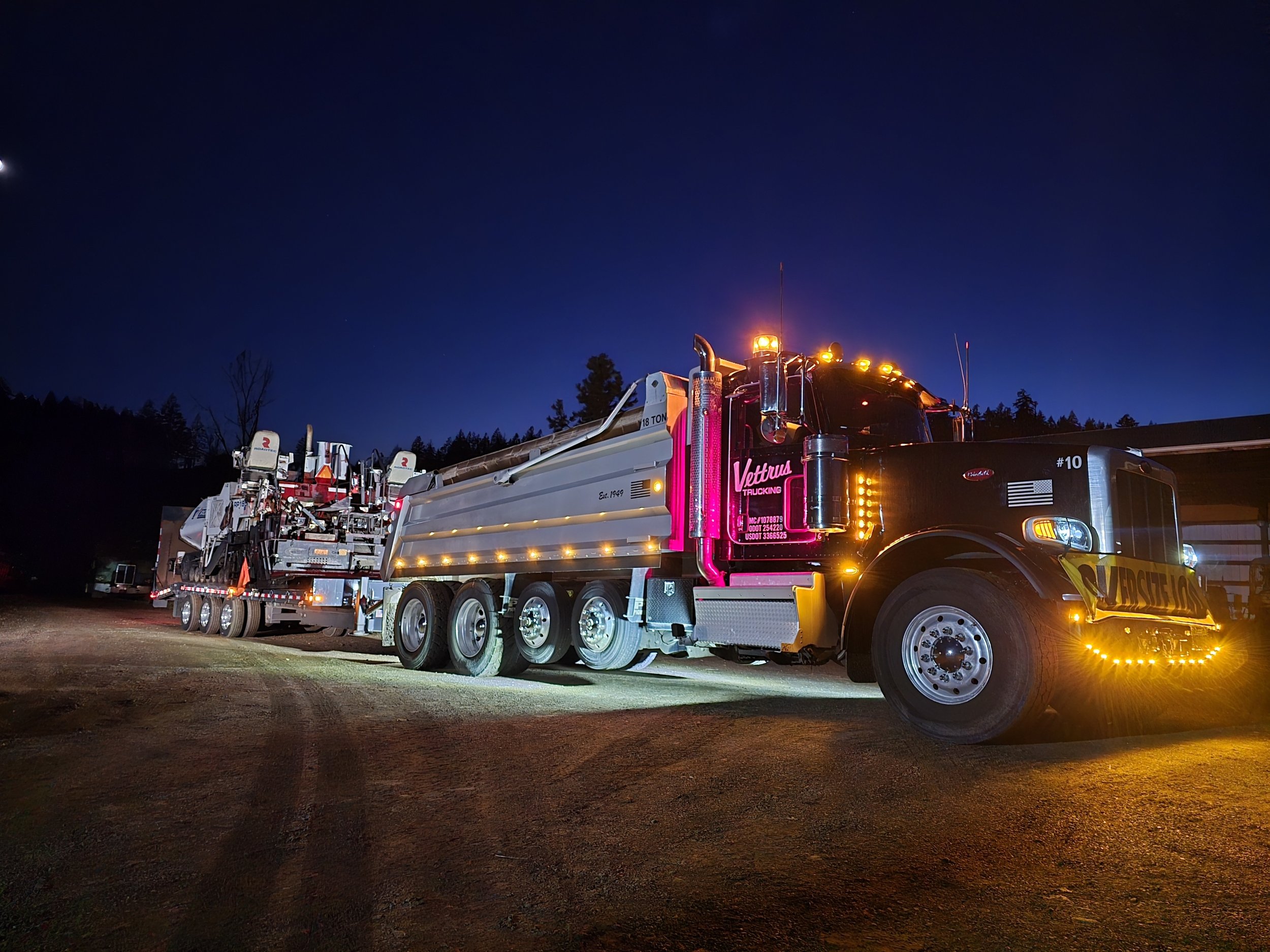 Nighttime scene of a large black and pink semi-truck with lights, pulling a long flatbed trailer carrying multiple boats with motors, parked on a dirt lot with trees in the background.