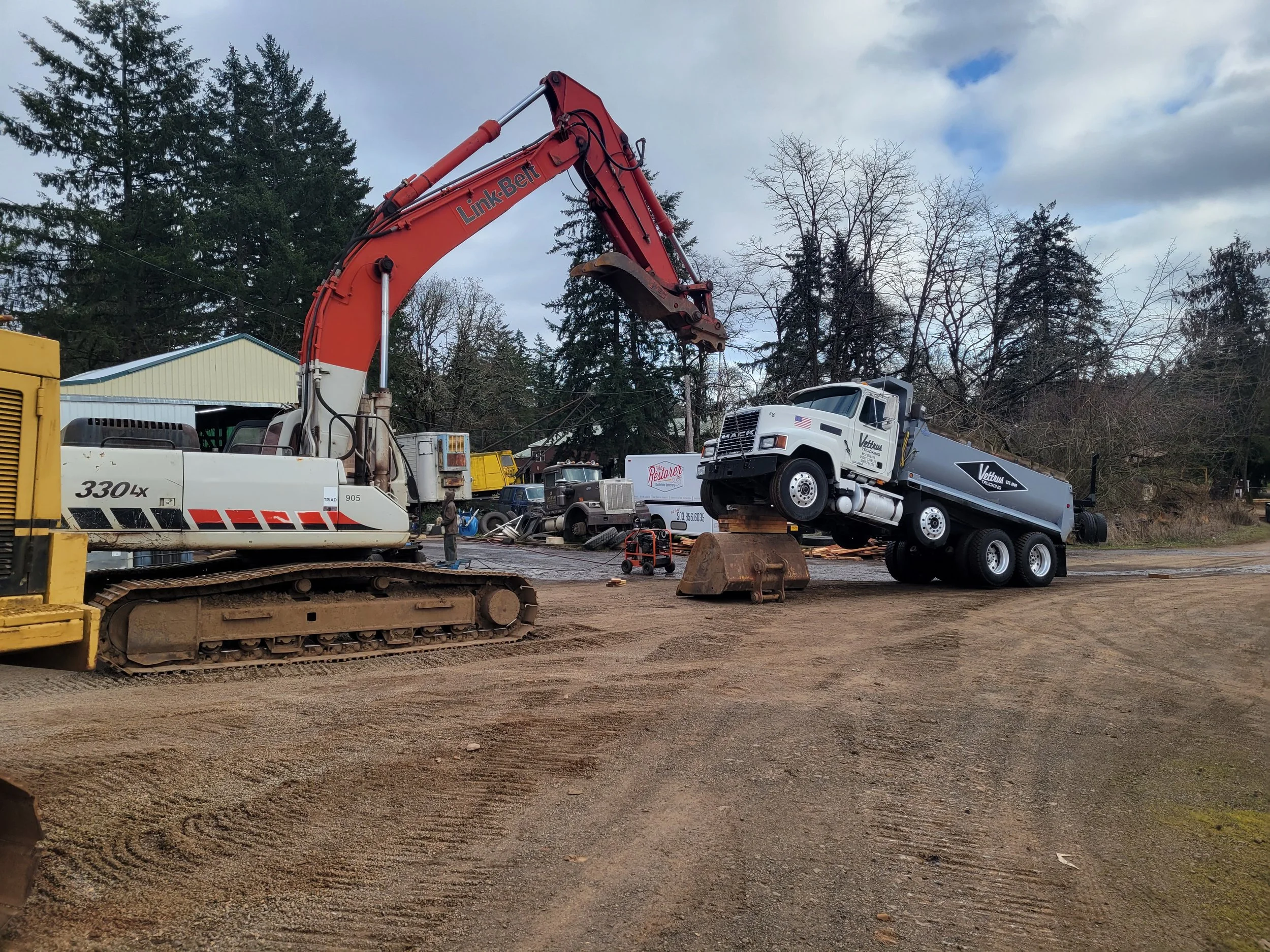 Construction yard with various heavy machinery, including a large orange excavator labeled Link-Belt lifting a white truck that is tilted backwards, surrounded by trees and a cloudy sky.