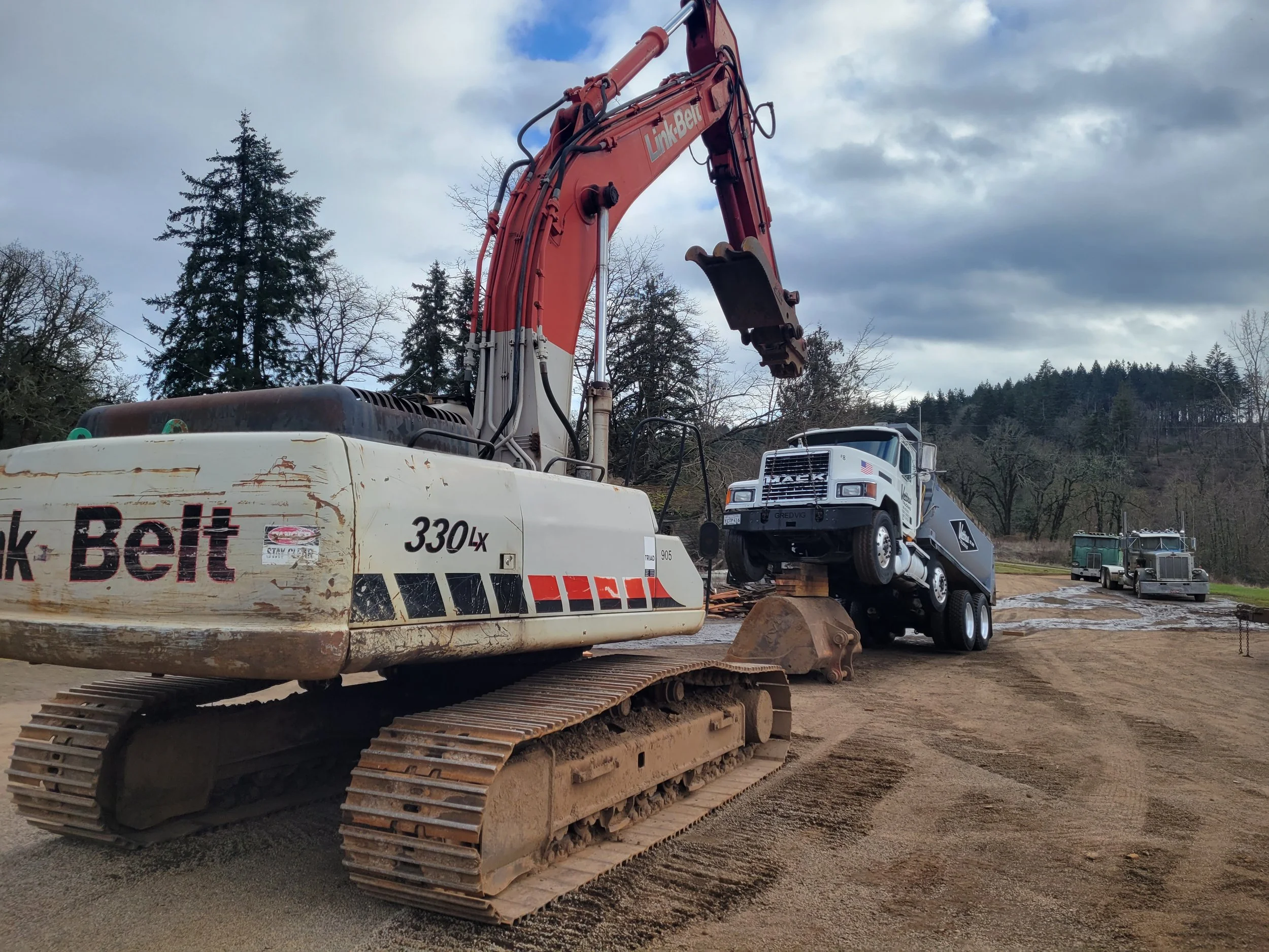 An excavator with a tracked undercarriage and a long arm reaching upward, parked on a construction site with dirt ground, trees in the background, and a partly cloudy sky overhead.