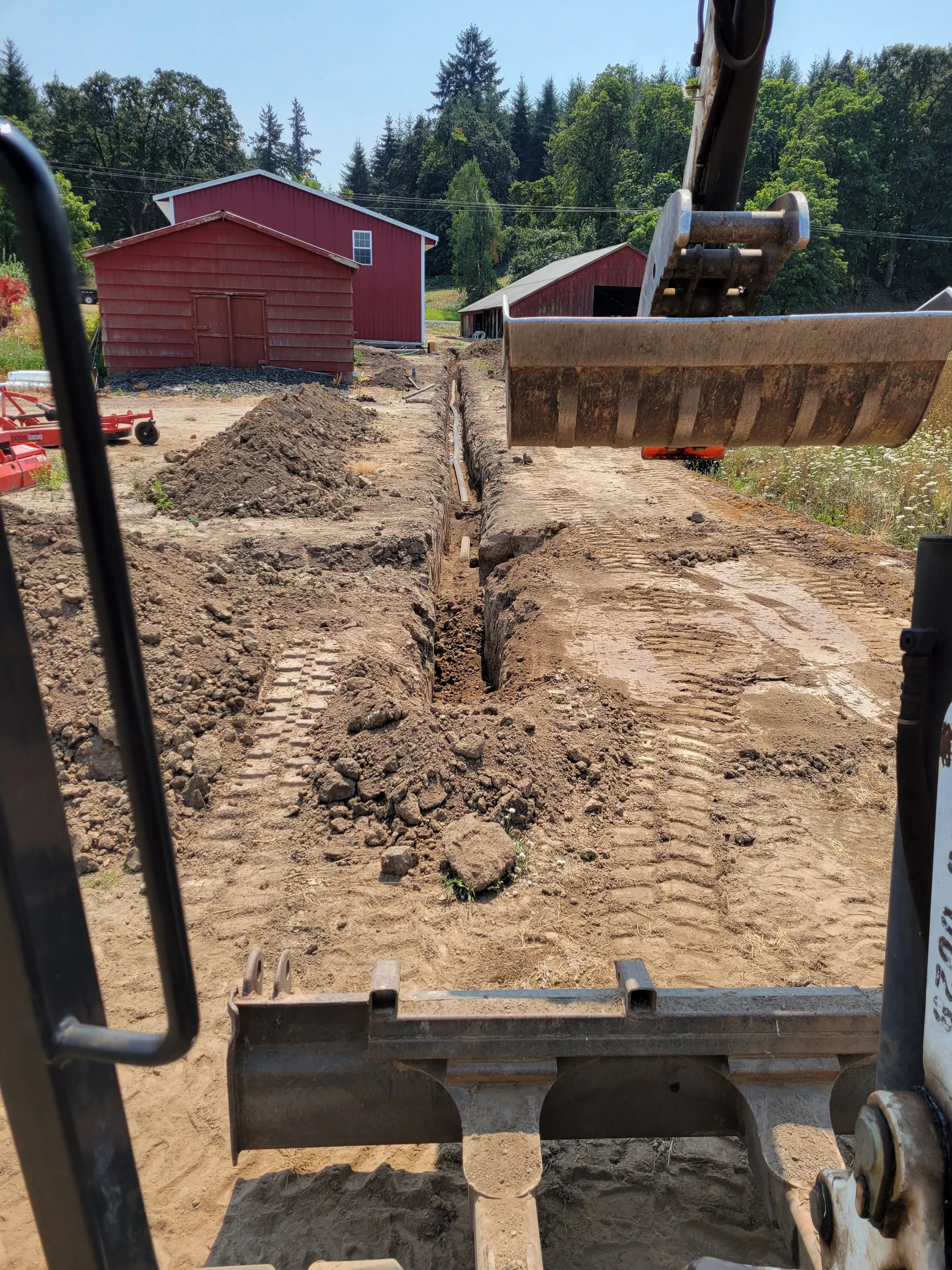 View from inside a construction vehicle showing a dirt road with a deep trench being dug, with red barns and trees in the background.