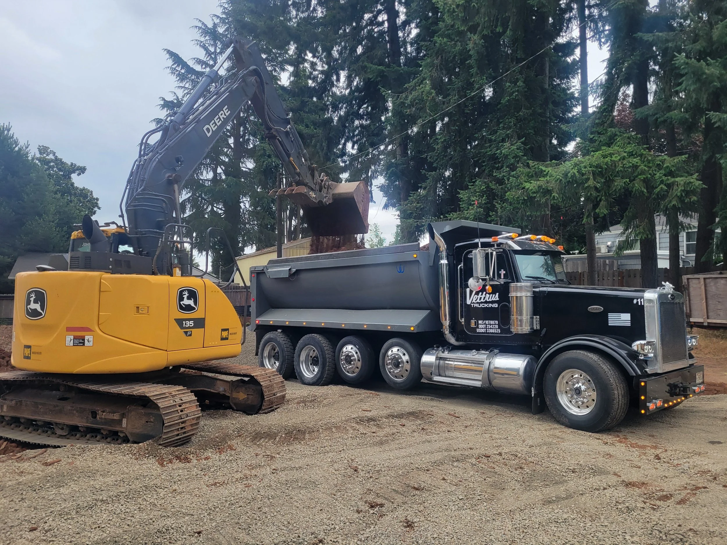 A construction site with a yellow John Deere excavator and a Vettrus truck. The excavator is lifting a large tree stump into the dump truck bed. In West Salem Oregon.