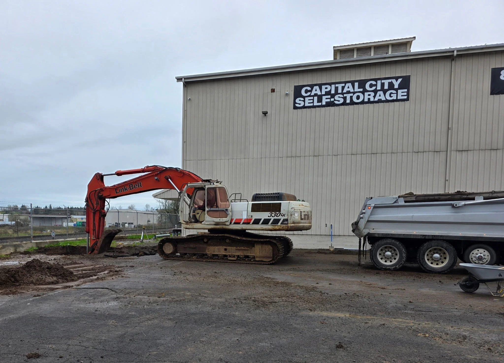 Construction site with an excavator and a truck in front of a building labeled 'Capital City Self-Storage' under an overcast sky. Turner Oregon.