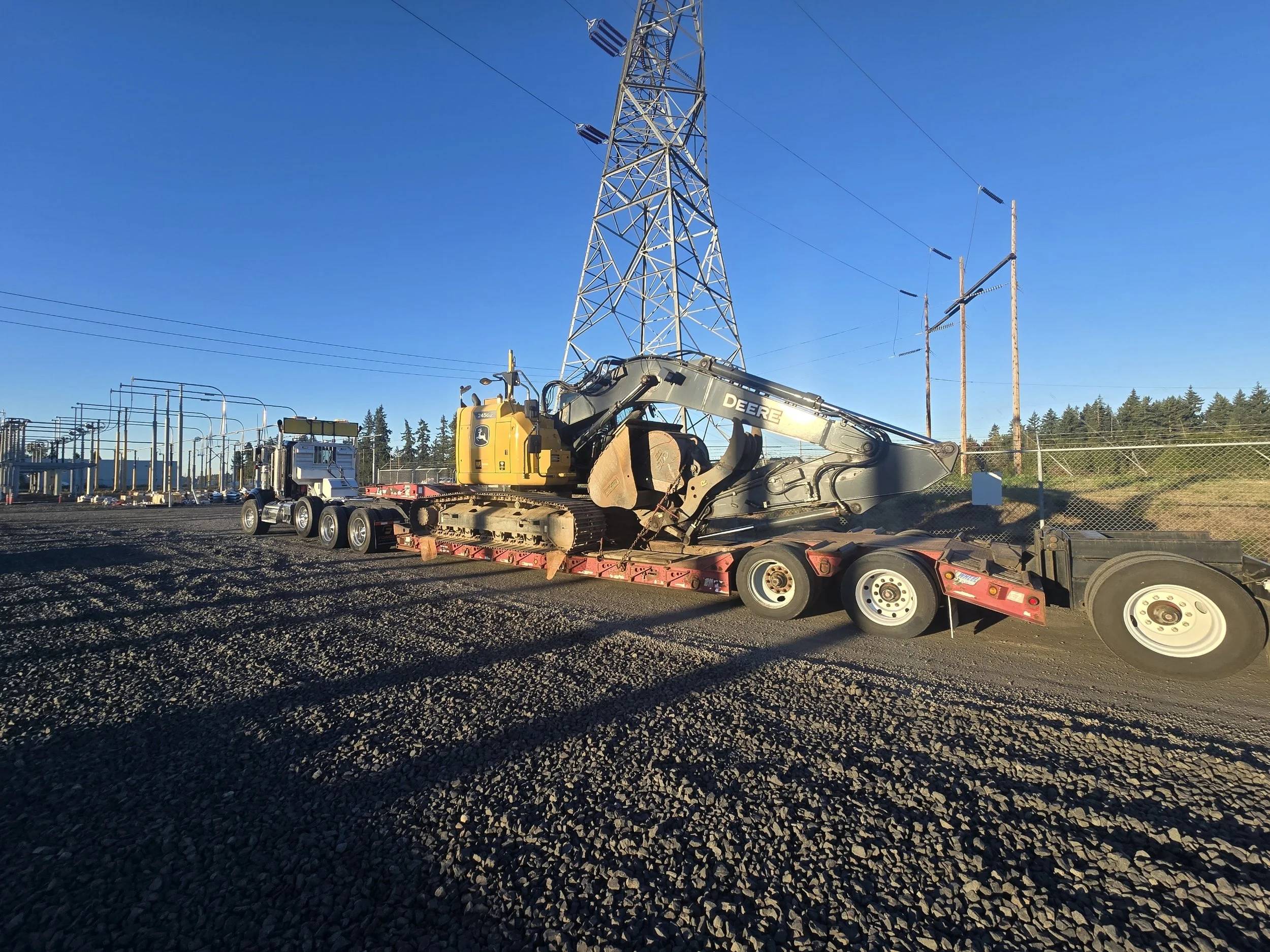 A construction site with a large electricity transmission tower and utility poles, with a yellow John Deere excavator on a trailer in the foreground, under a clear blue sky. Salem Oregon