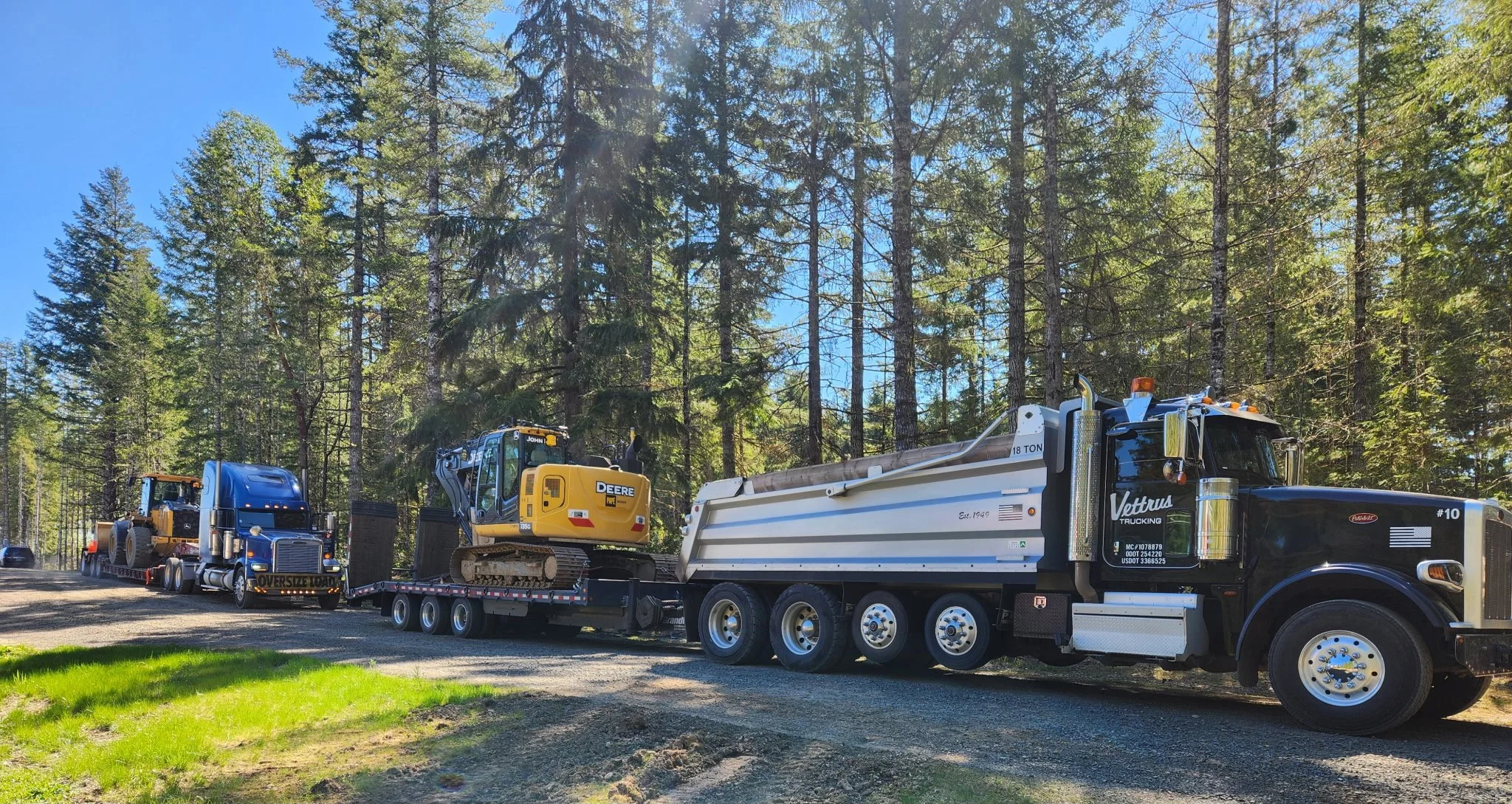 A large semi-truck hauling heavy equipment, including a yellow John Deere excavator, through a forested area with tall trees and bright blue sky.