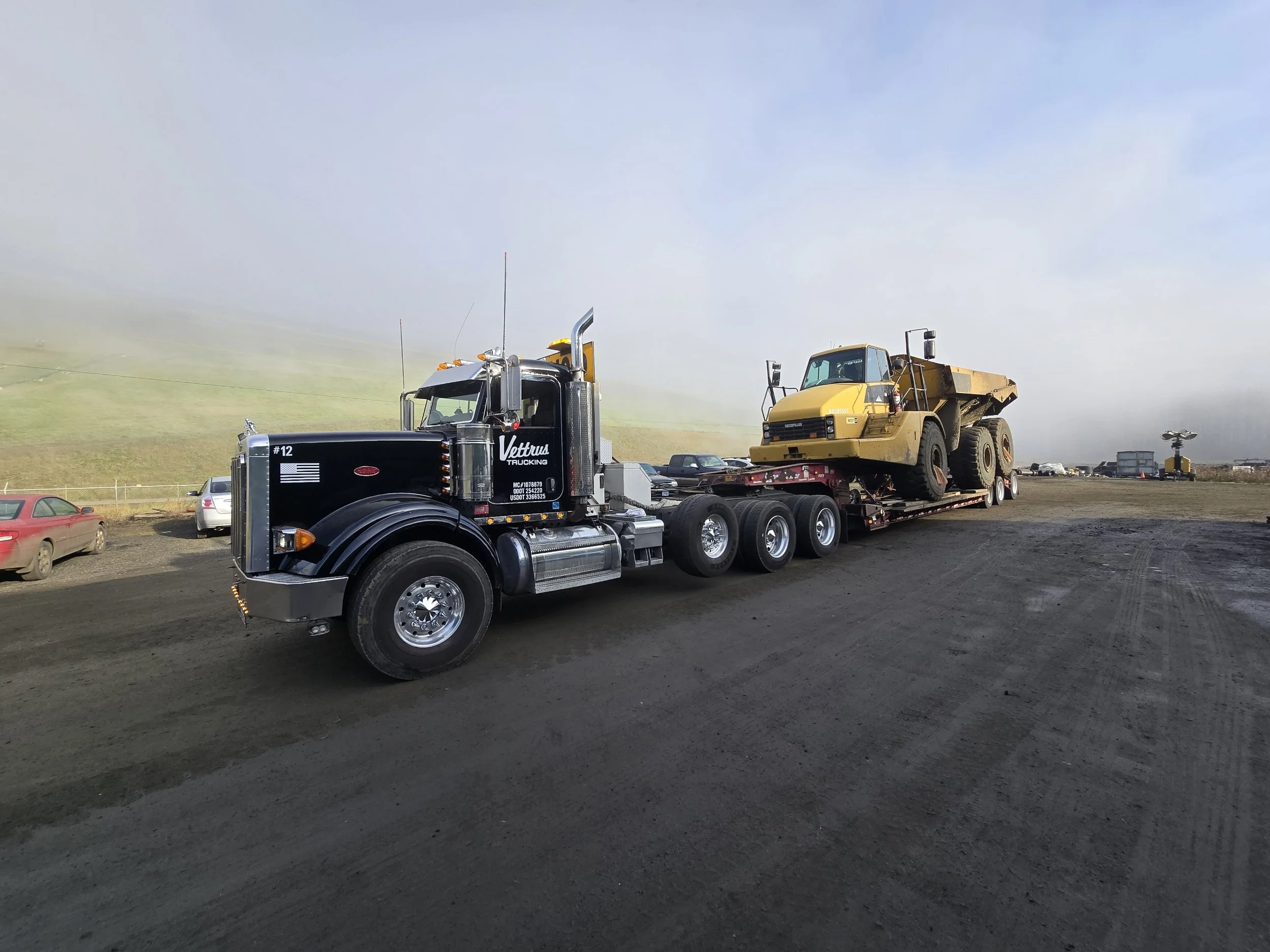 A black semi-truck carrying a yellow construction vehicle on a flatbed trailer parked on a dirt lot under a cloudy sky.