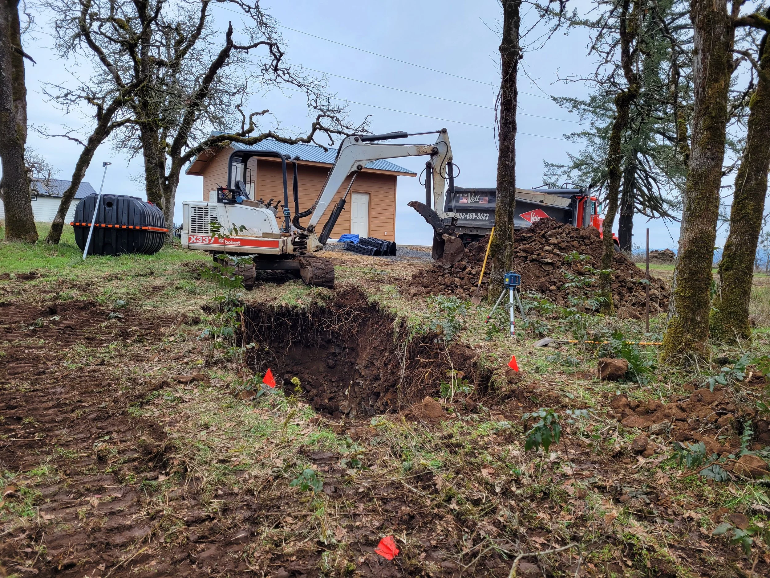 Construction site with a mini excavator digging a trench, piles of rocks, a black tank, and trees with moss in the background. Septic tank install Dallas Oregon