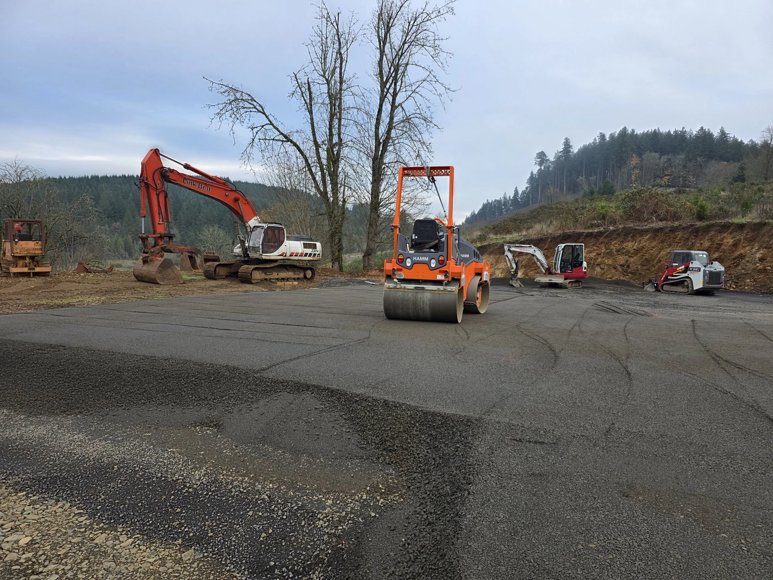 Construction equipment on a project site with trees and hills in the background, including an excavator, a compactor, and other machinery.