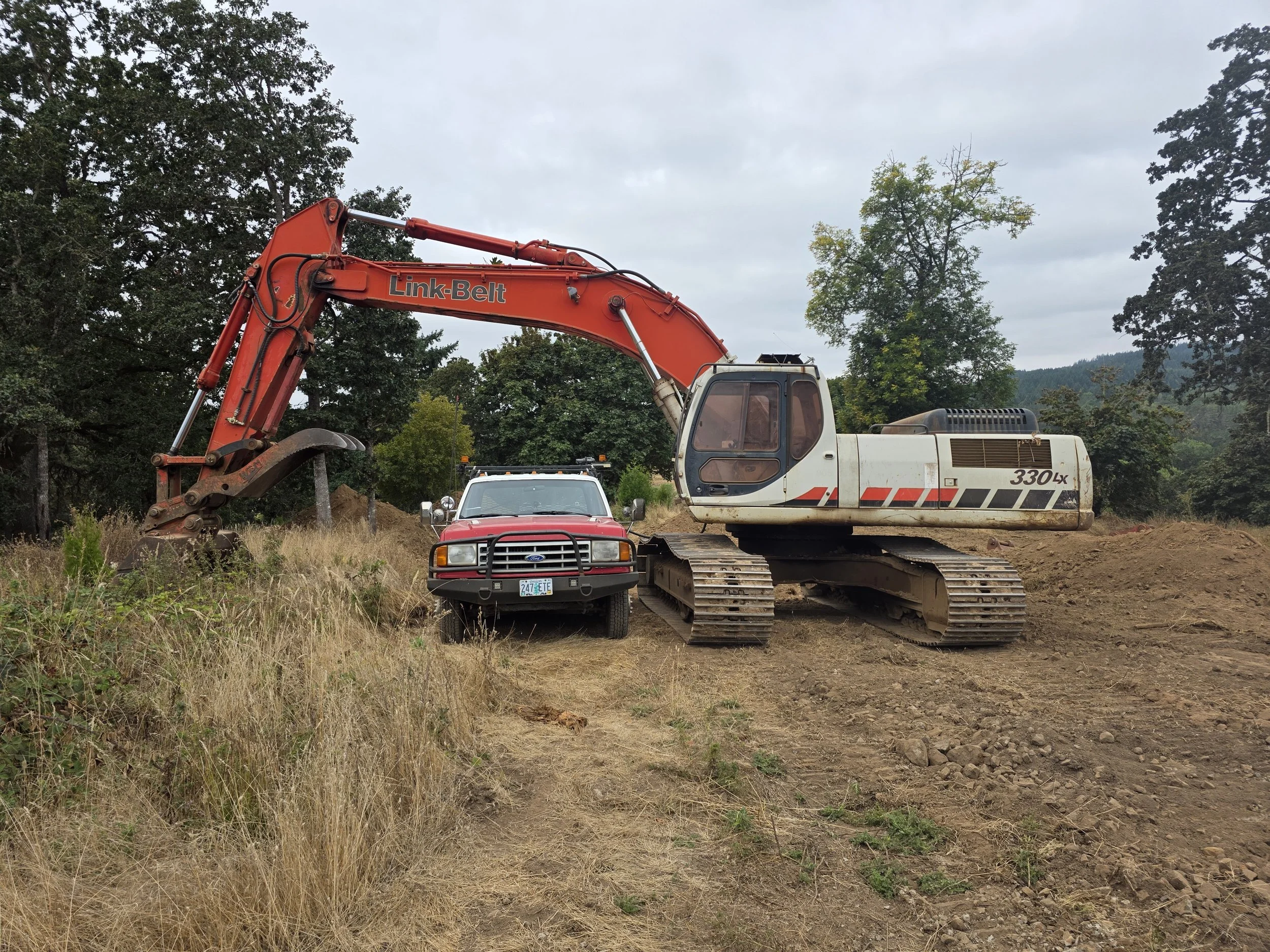 A construction site with an orange excavator labeled 'Link-Belt' parked next to a white pickup truck and a red Ford vehicle. The area has dirt and grass, with trees and a cloudy sky in the background.