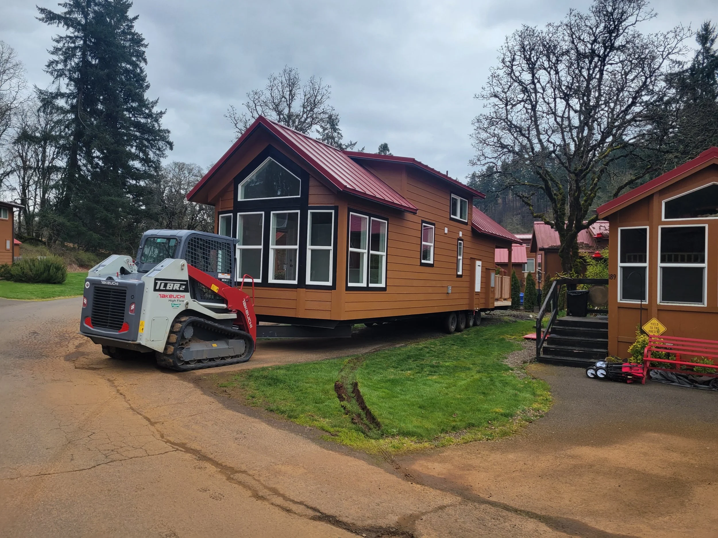 A tiny house on a trailer being moved by a small construction vehicle, located on a residential street with trees and other houses.