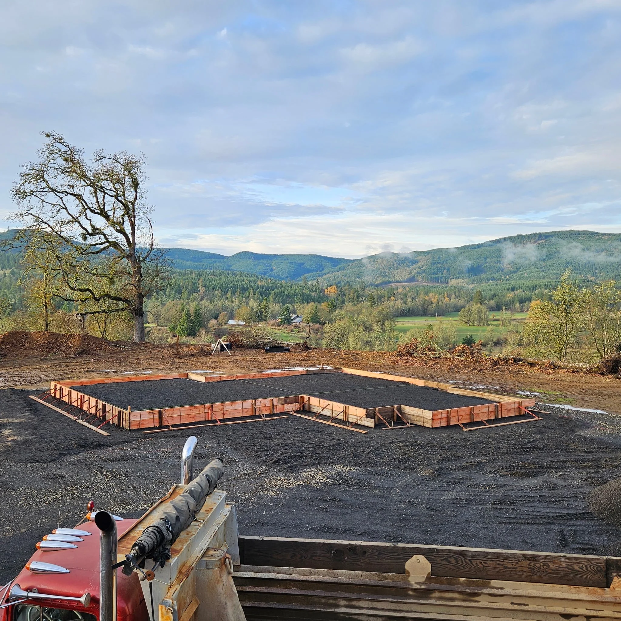 Construction site with a concrete foundation in progress, surrounded by a rural landscape with hills and trees, and a cloudy sky. Brownsville Oregon.